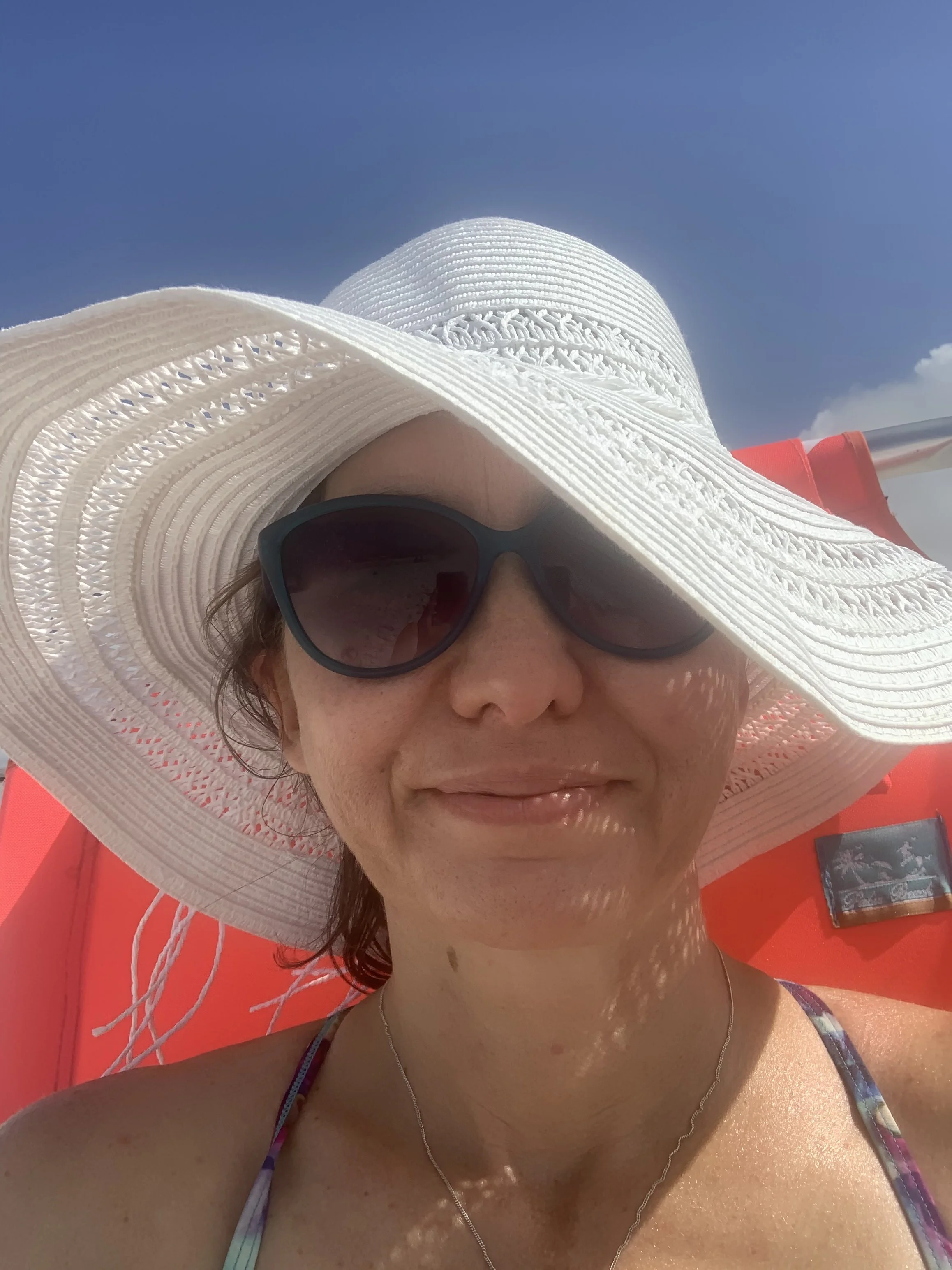 A woman wearing sunglasses and a large white sunhat, with a red background, possibly on a boat or outdoor setting.
