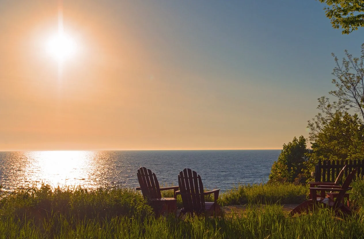 Sunset over the ocean viewed from a grassy area with four wooden chairs and a fence on the right, surrounded by green trees.