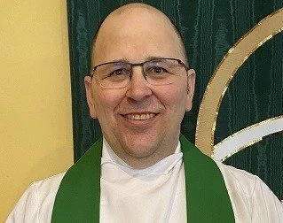 A smiling man with glasses, wearing a white shirt and a green stole, standing indoors with a green and yellow background.