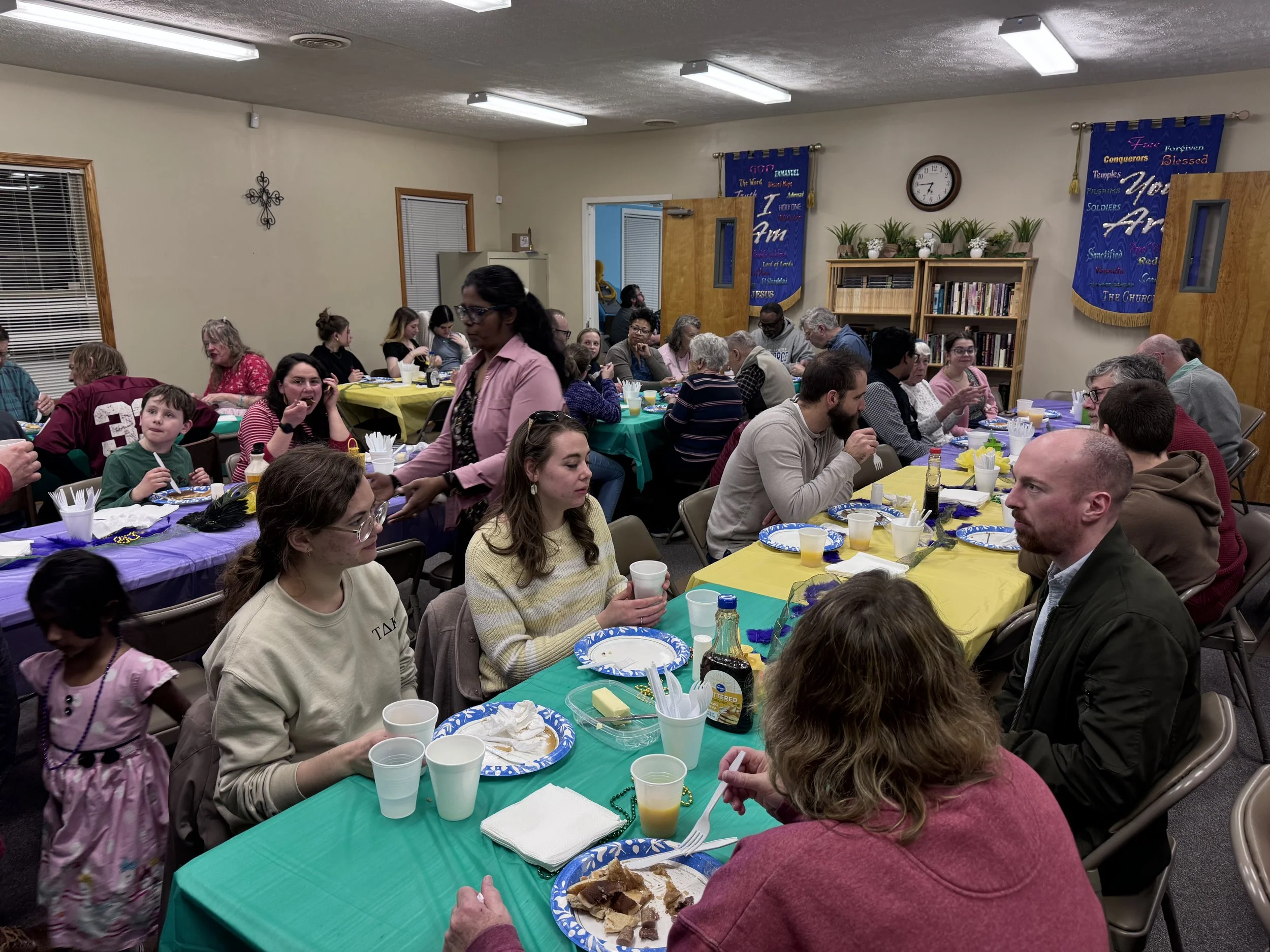 A large group of people gathered in a room with decorated tables, enjoying a meal together, with banners hanging on the walls and a clock showing the time.