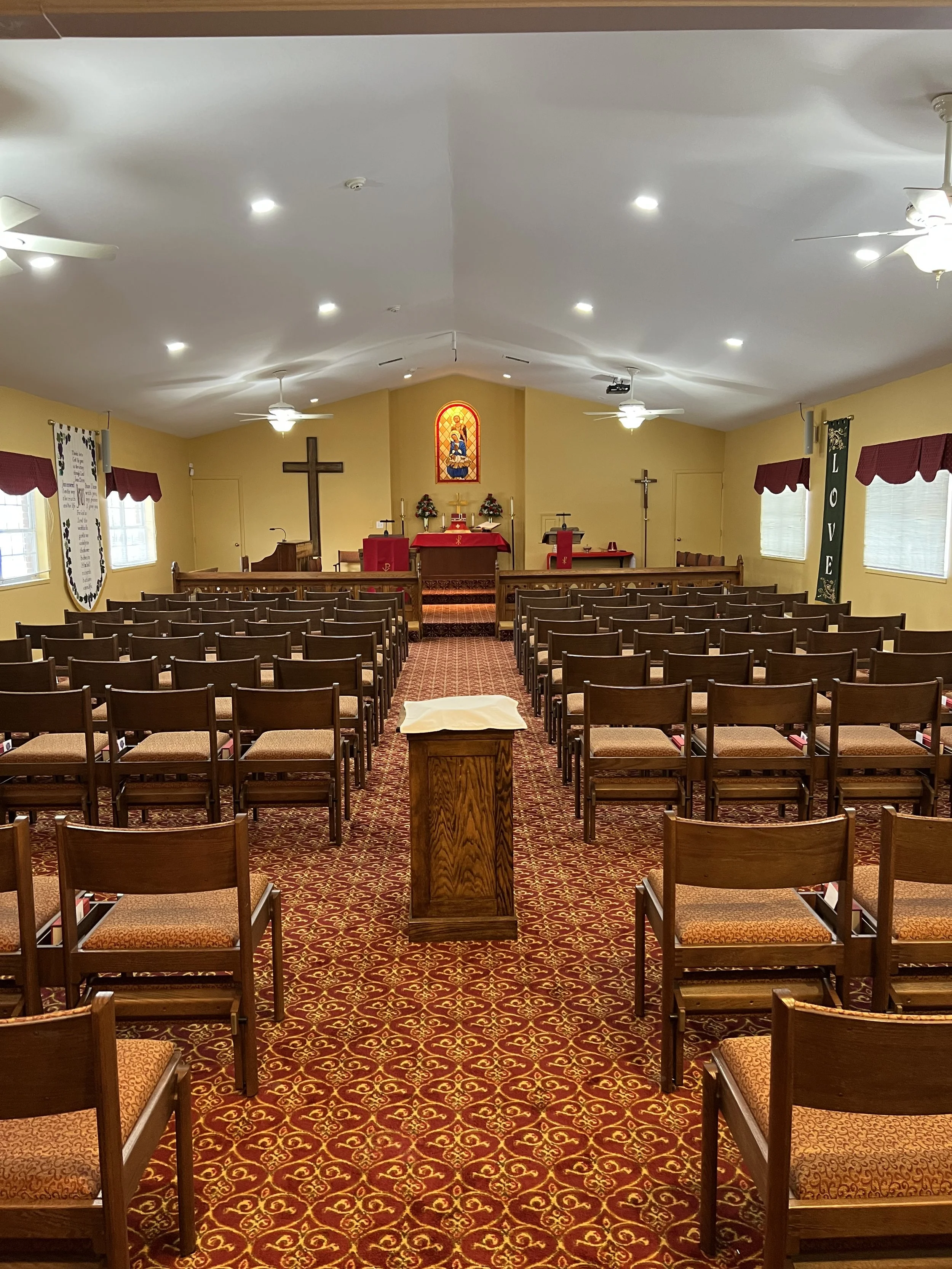 Inside a church with wooden chairs arranged in rows, a wooden podium at front on a patterned carpet, a red altar cloth and candles, religious banners on the walls, stained glass window depicting a saint, crosses on the walls, and ceiling fans with lights.