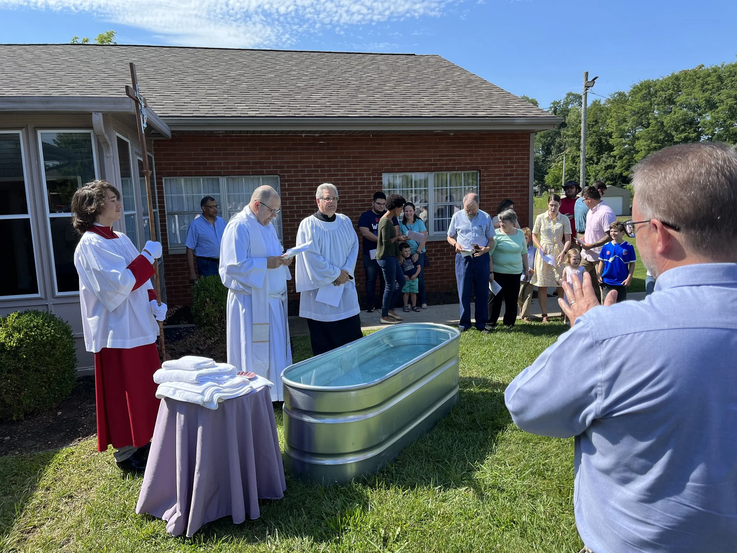A group of people, including a priest and choir members, gathered outside a house for a baptism ceremony. There is a water baptism font and towels on a table, with some attendees holding papers.