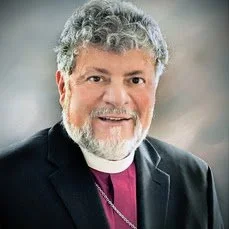 A middle-aged man with gray hair and a beard, wearing clerical clothing and a necklace, smiling indoors.