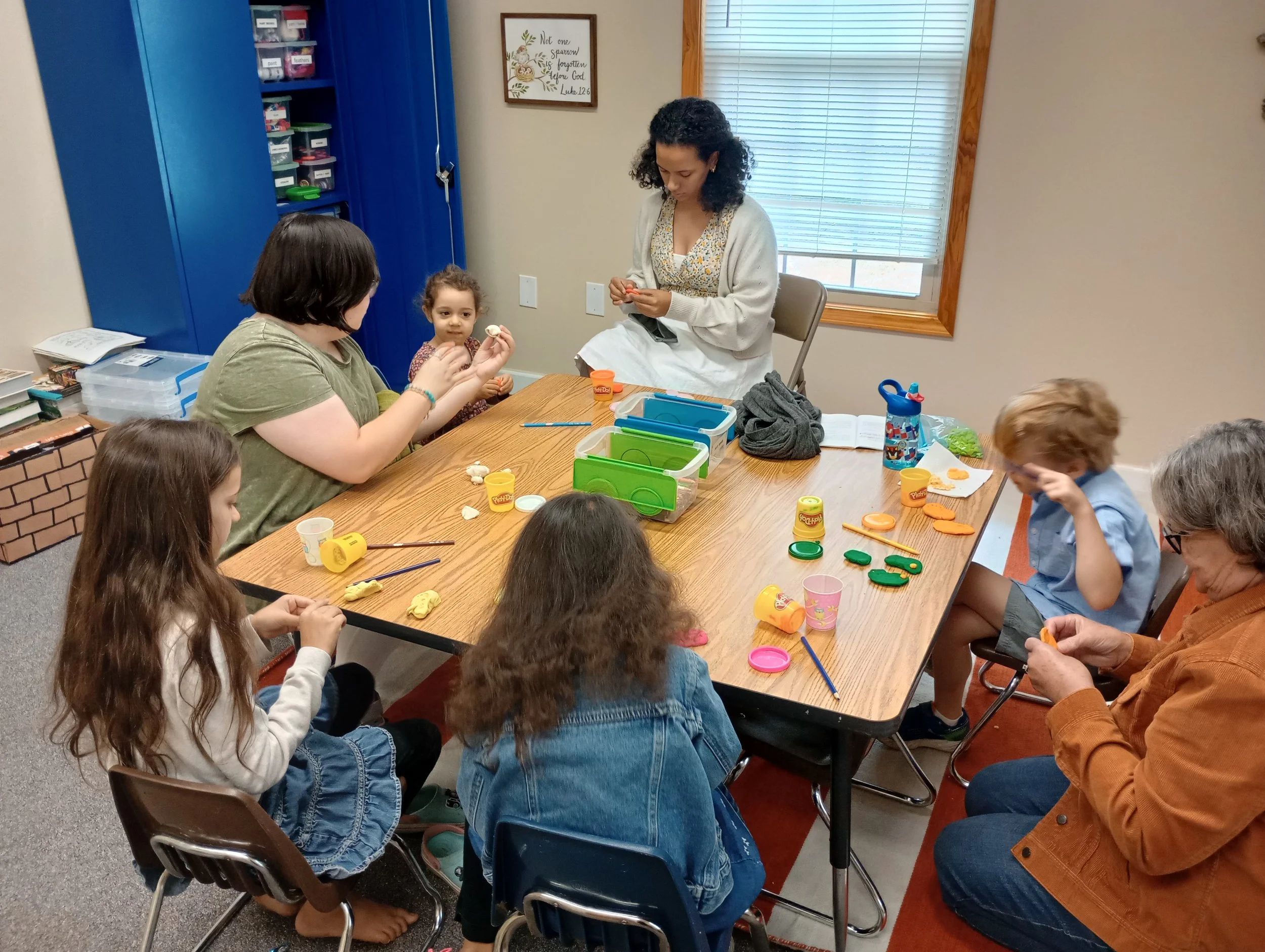 A group of children and an adult woman sitting around a table in a classroom, engaging in arts and crafts activities with Play-Doh and other supplies.