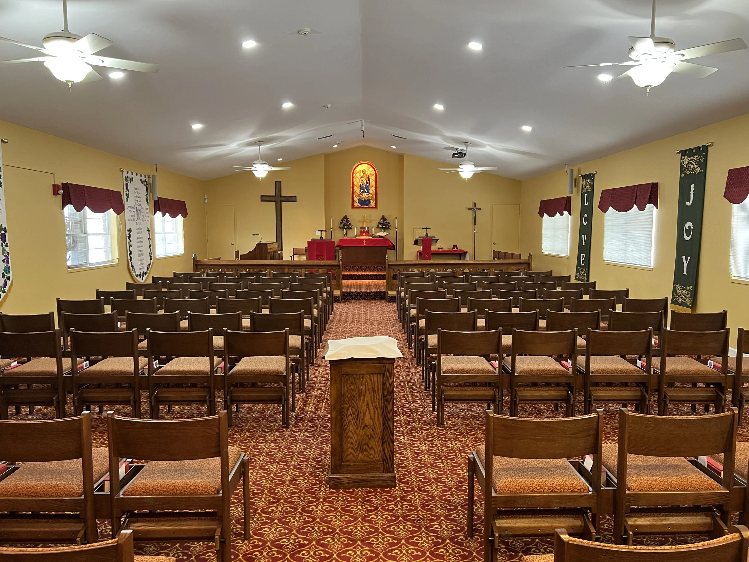 Interior of a church sanctuary with rows of wooden pews, a central aisle, an altar at the front, crosses, stained glass window, and religious banners, with ceiling fans and lights.