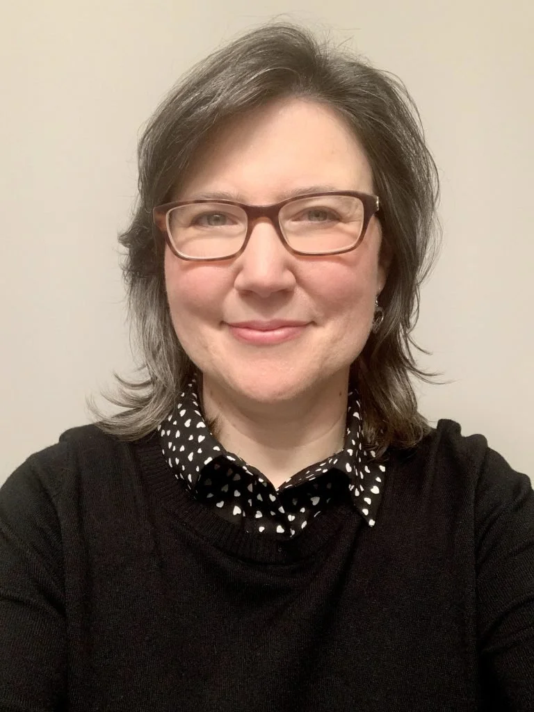 A woman with shoulder-length gray hair, wearing glasses, a black sweater, and a black and white polka dot collared shirt, smiling at the camera against a plain beige wall.