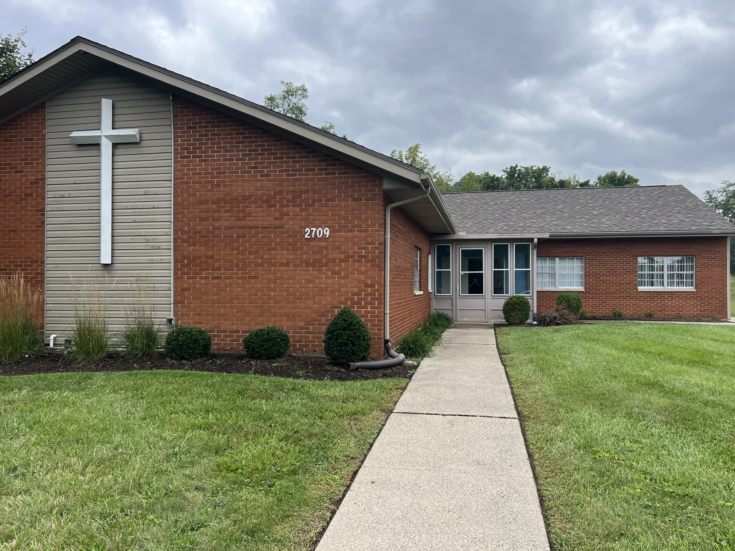 An exterior view of a brick church building with a large metal cross on the front, multiple windows, a small landscaped yard, and a concrete walkway leading to the entrance, under a cloudy sky.