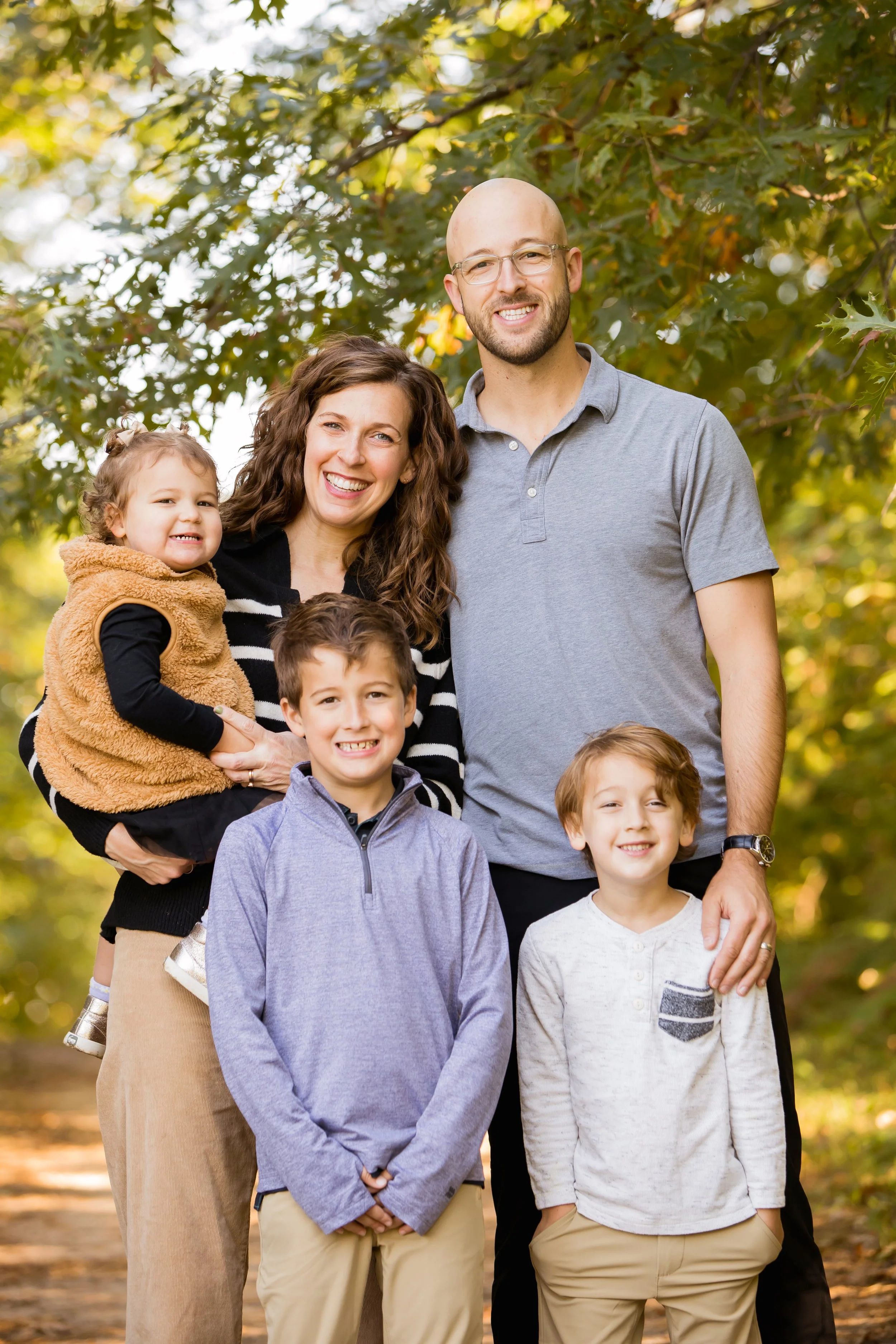 Family of seven smiling outdoors in fall, with trees and sunlight in the background.