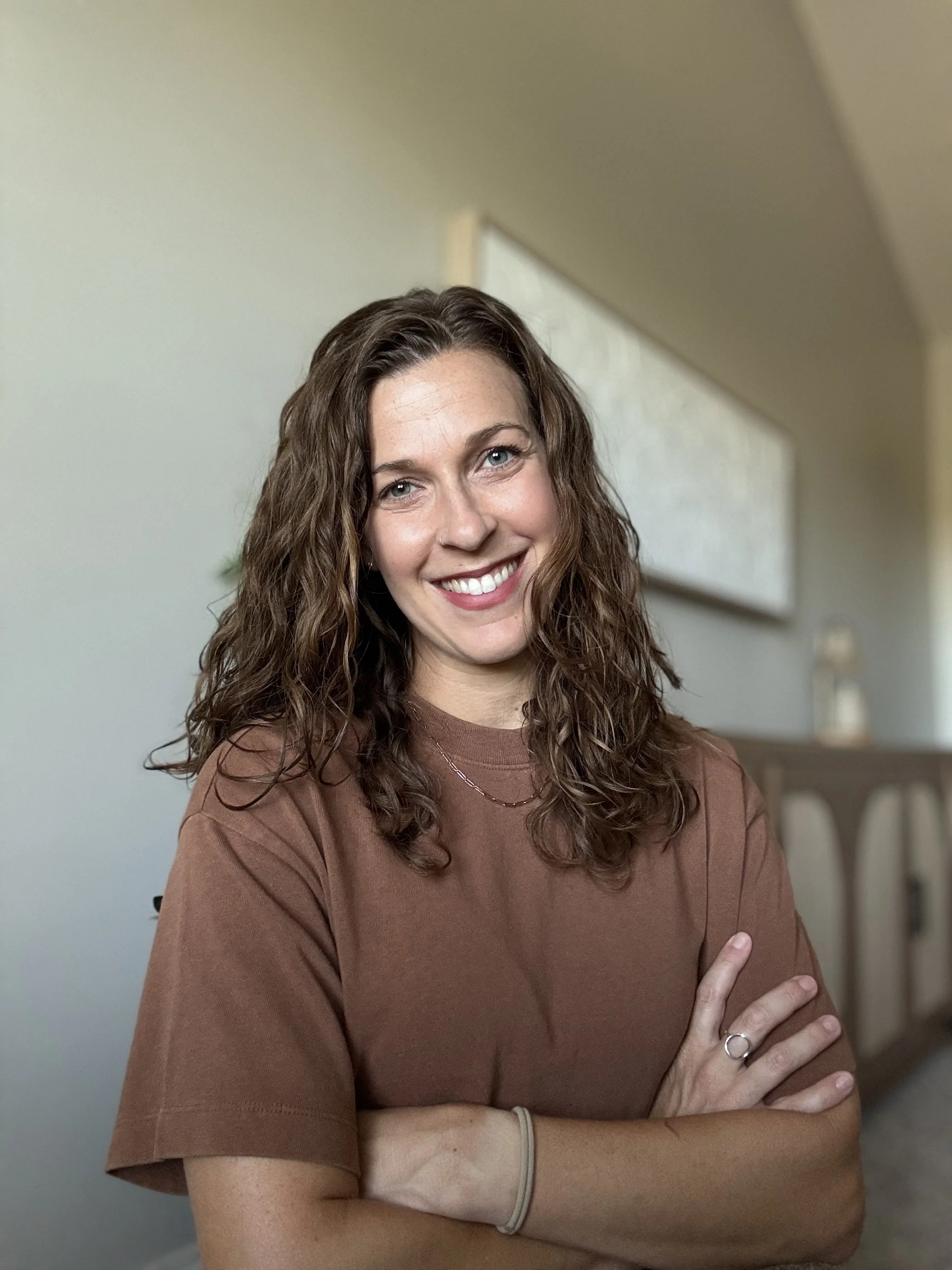 A young woman with curly brown hair, light blue eyes, and fair skin, smiling and looking at the camera, standing in a room with a neutral-colored wall and furniture in the background.
