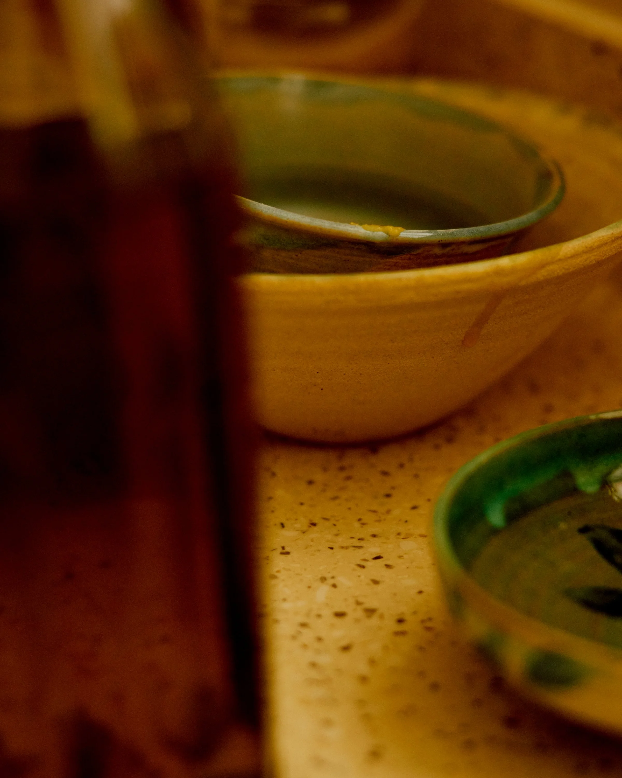 Close-up of a yellow ceramic cup and a green ceramic cup on a speckled beige surface, with a partial view of a copper-colored object and a metallic spoon in the background.