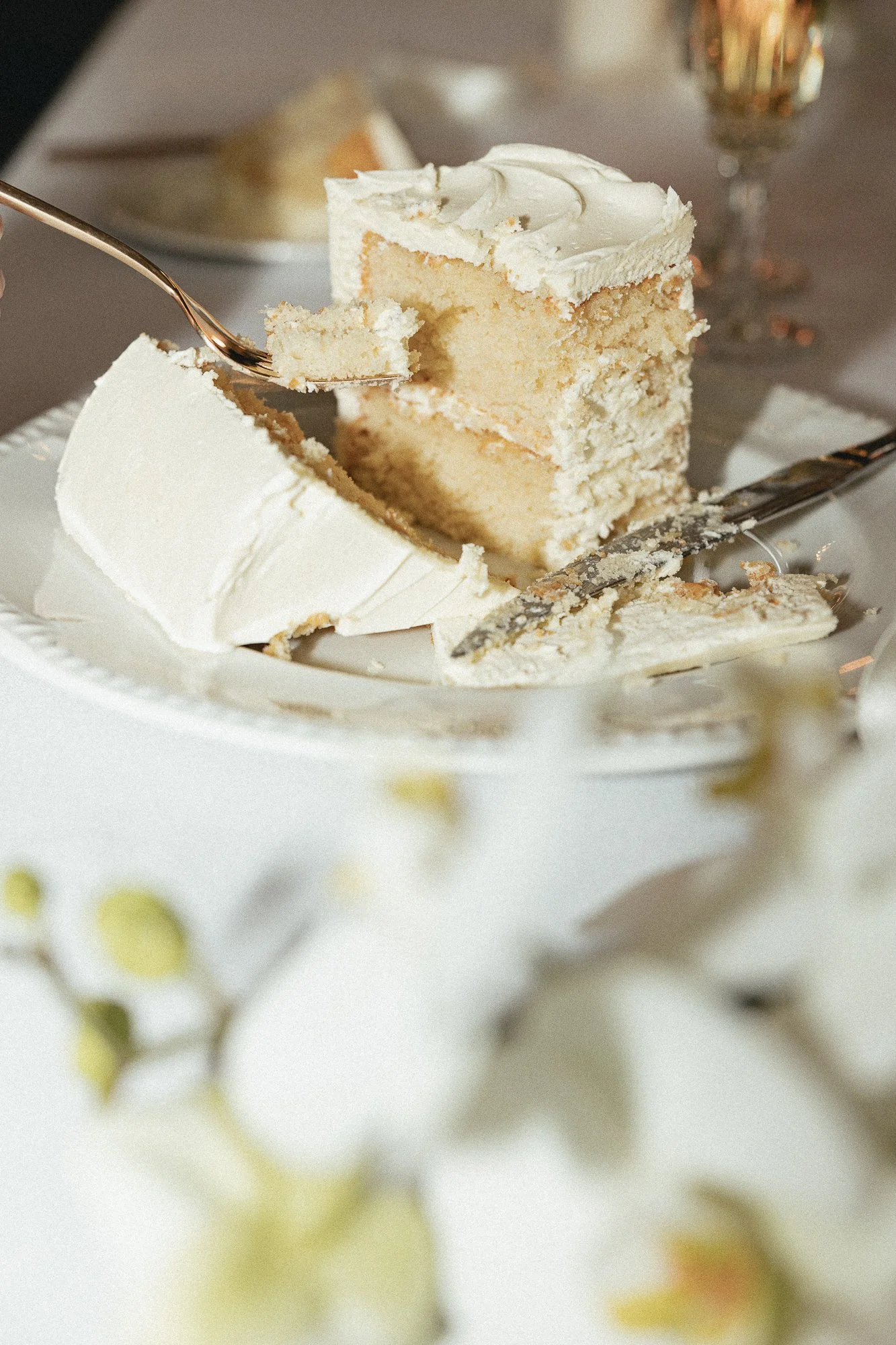 A slice of vanilla cake with white frosting on a white plate, with a fork and knife.