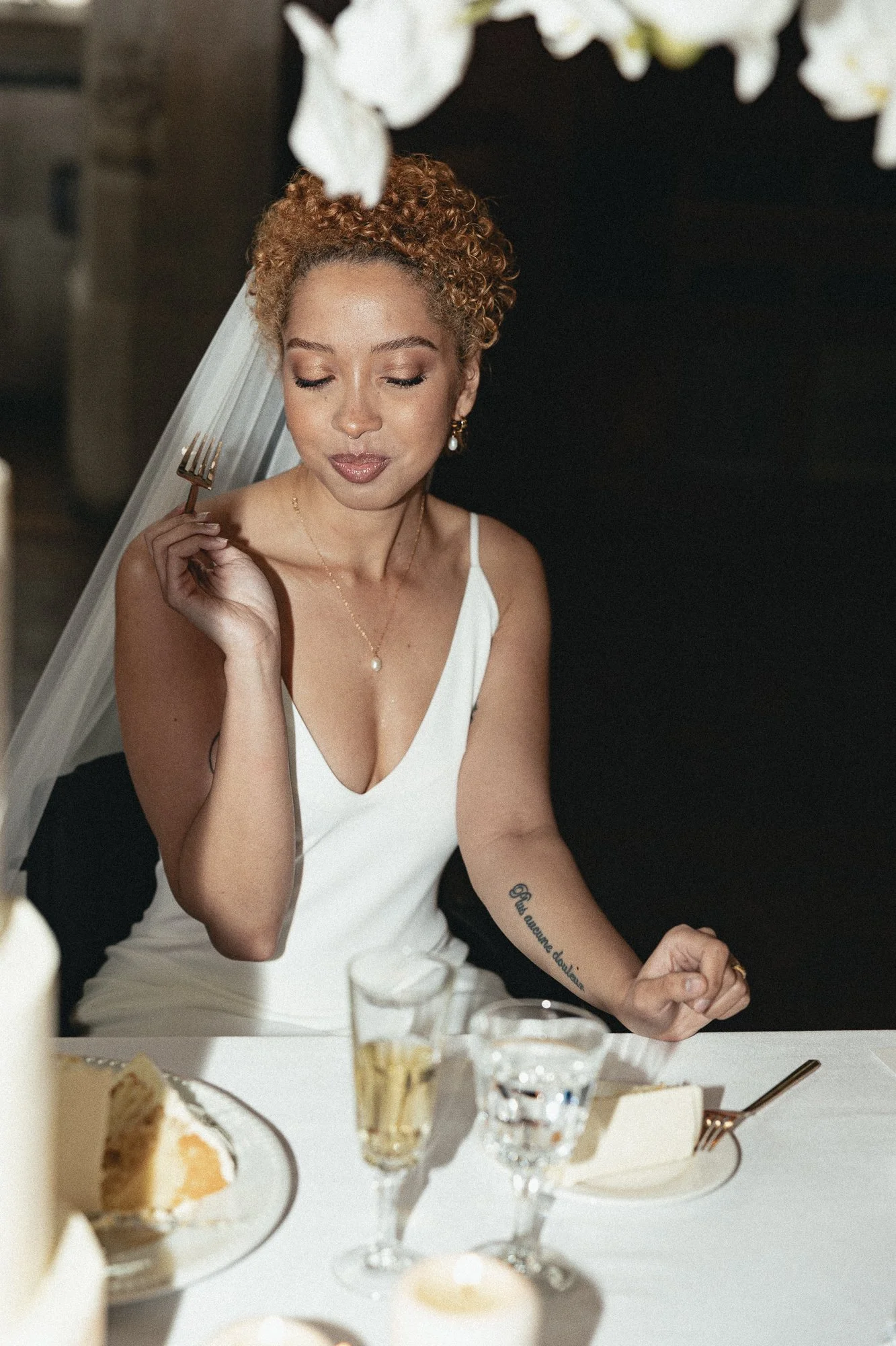 A woman with curly hair in a white dress is sitting at a dinner table, holding a fork near her face with her eyes closed. The table is decorated with food, desserts, glasses, and candles, with a dark backdrop.