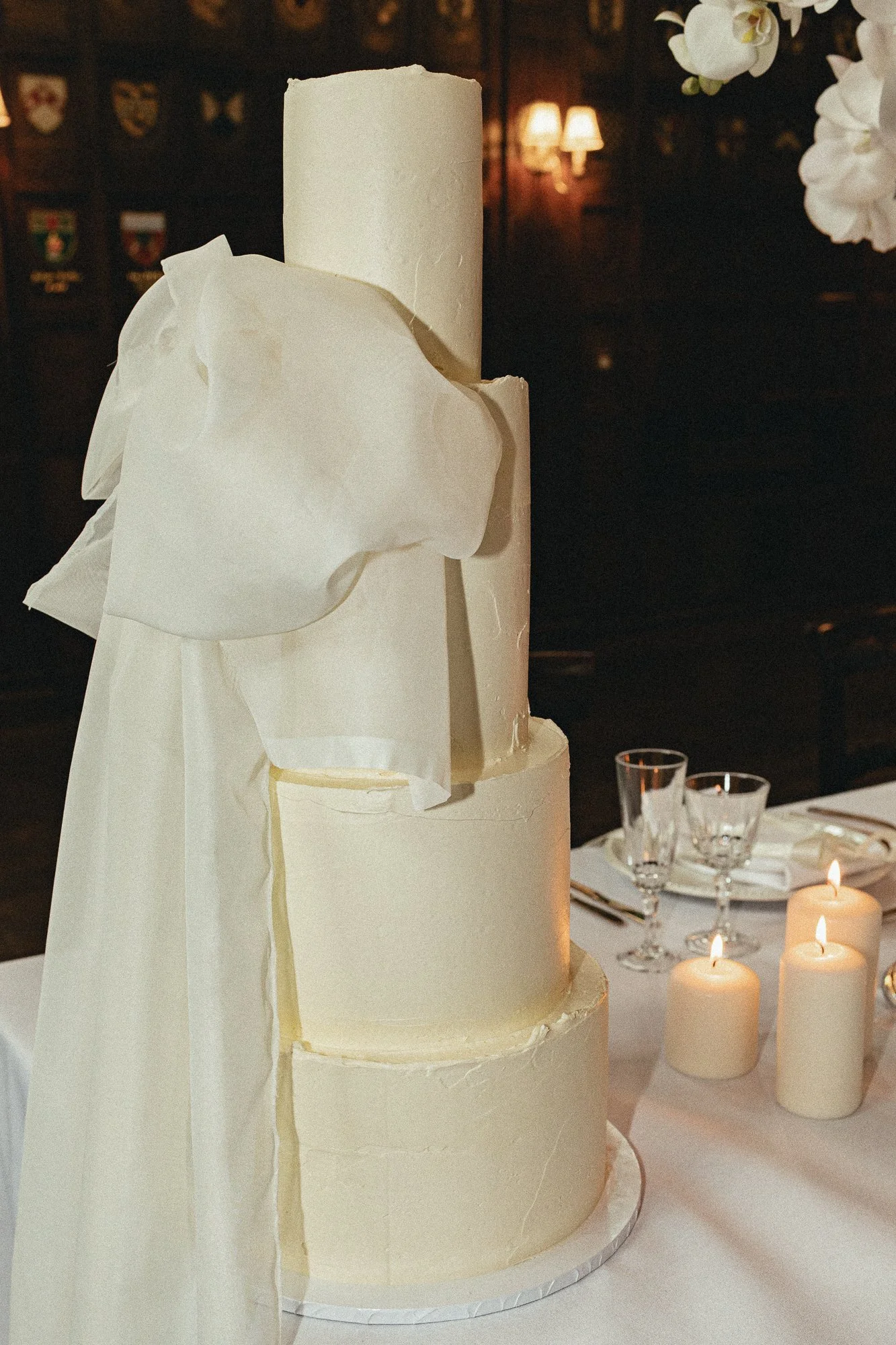 A multi-tiered white wedding cake with a minimalist design, decorated with white edible flower accents, set on a table with lit candles and glassware in the background.
