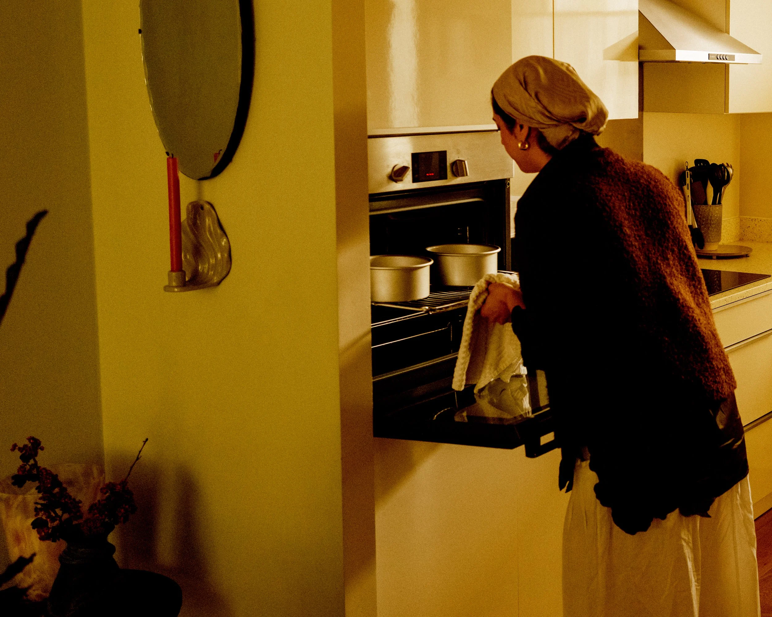 A woman with blonde hair in a bun, wearing earrings, a purple sweater, and beige pants, is standing in a kitchen, looking into an oven while holding a dish towel. The kitchen has cream-colored cabinets, a yellow wall, a stove, and a utensil holder. T