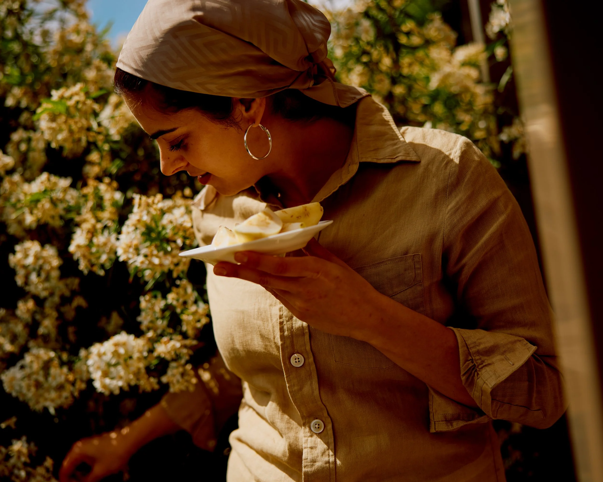 A woman wearing hoop earrings and a beige headscarf, looking downward, holding a white plate with sliced bananas, outdoors with yellow flowers and greenery in the background.