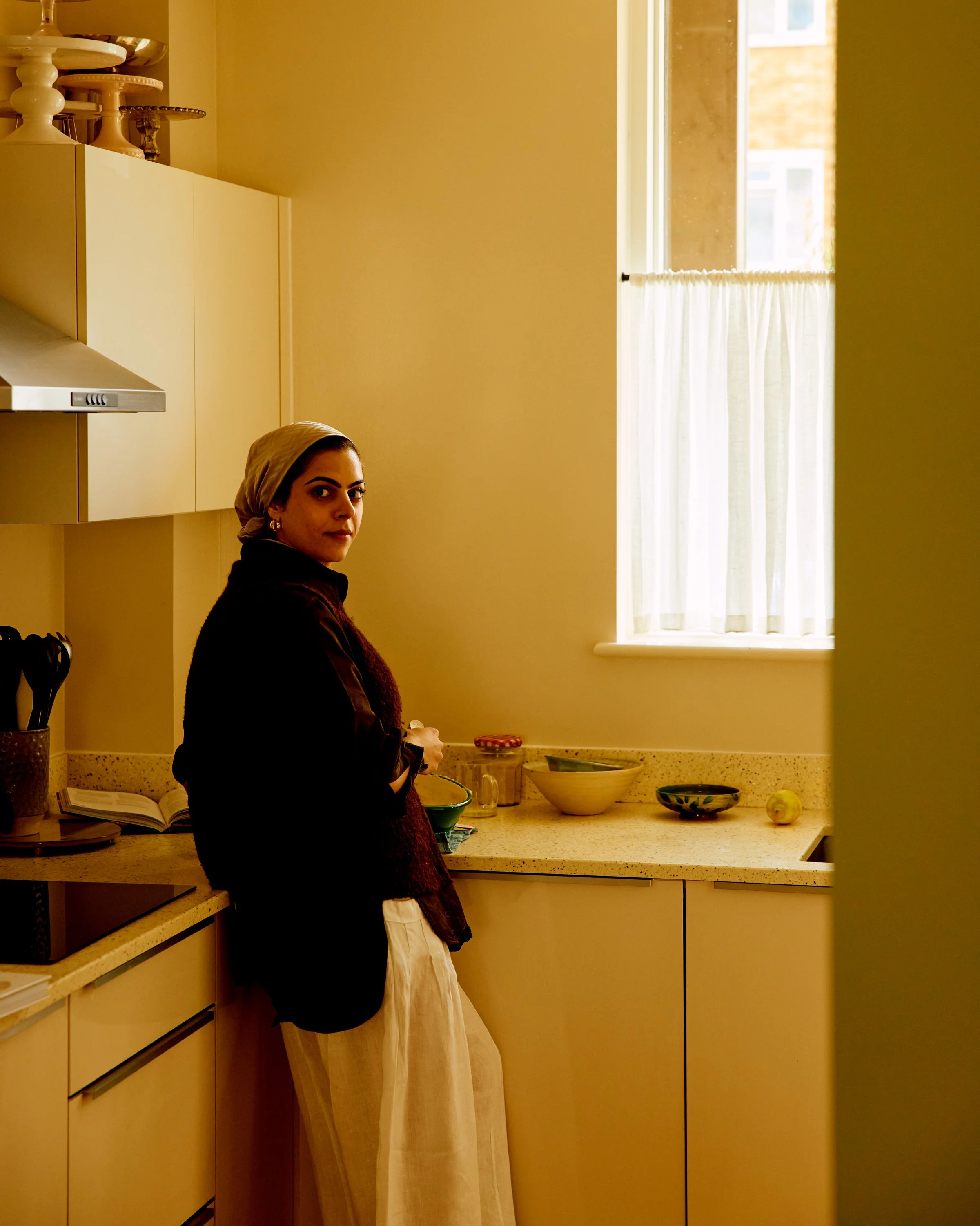 Woman with dark hair and a headscarf standing in a kitchen with yellow walls, white cabinets, and a window with white curtains, looking towards the camera.