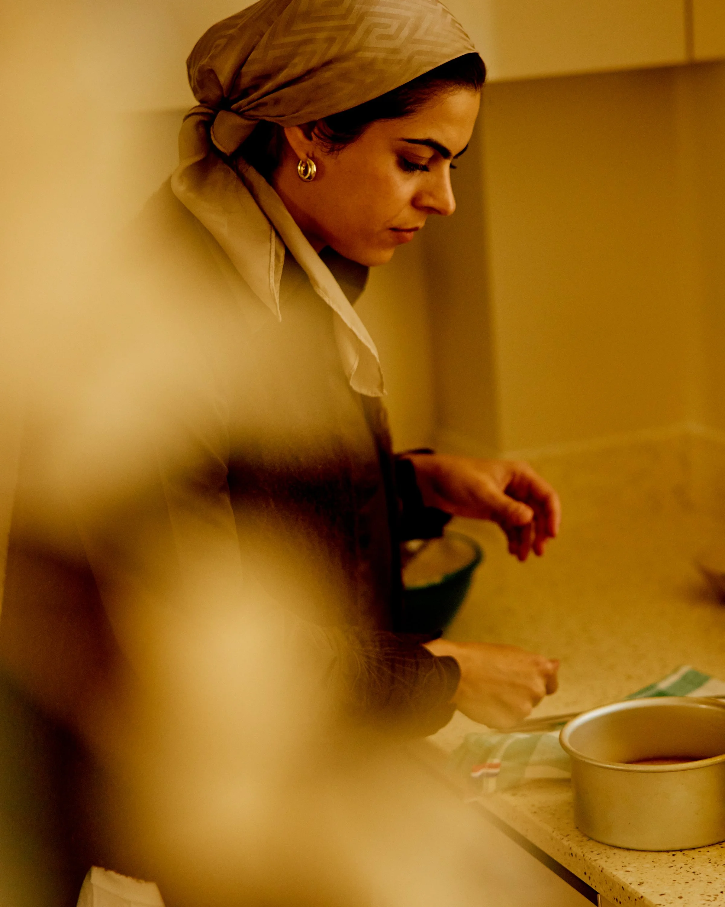A woman wearing a headscarf and earrings looks down while standing in a kitchen. There is a large white container on the countertop and a bowl of colorful objects.