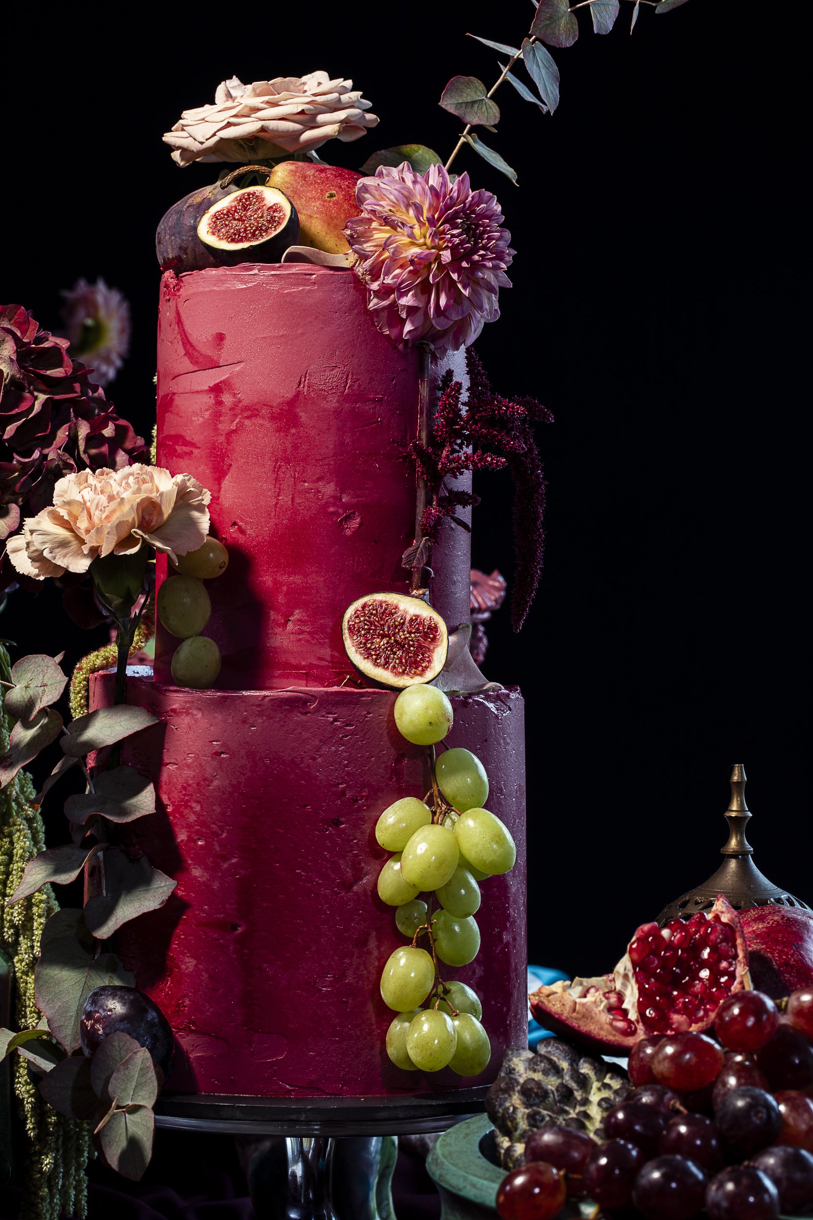 A pink multi-tiered cake decorated with fruits like figs, grapes, and flowers, with additional fruits and desserts on a plate nearby, against a dark background.