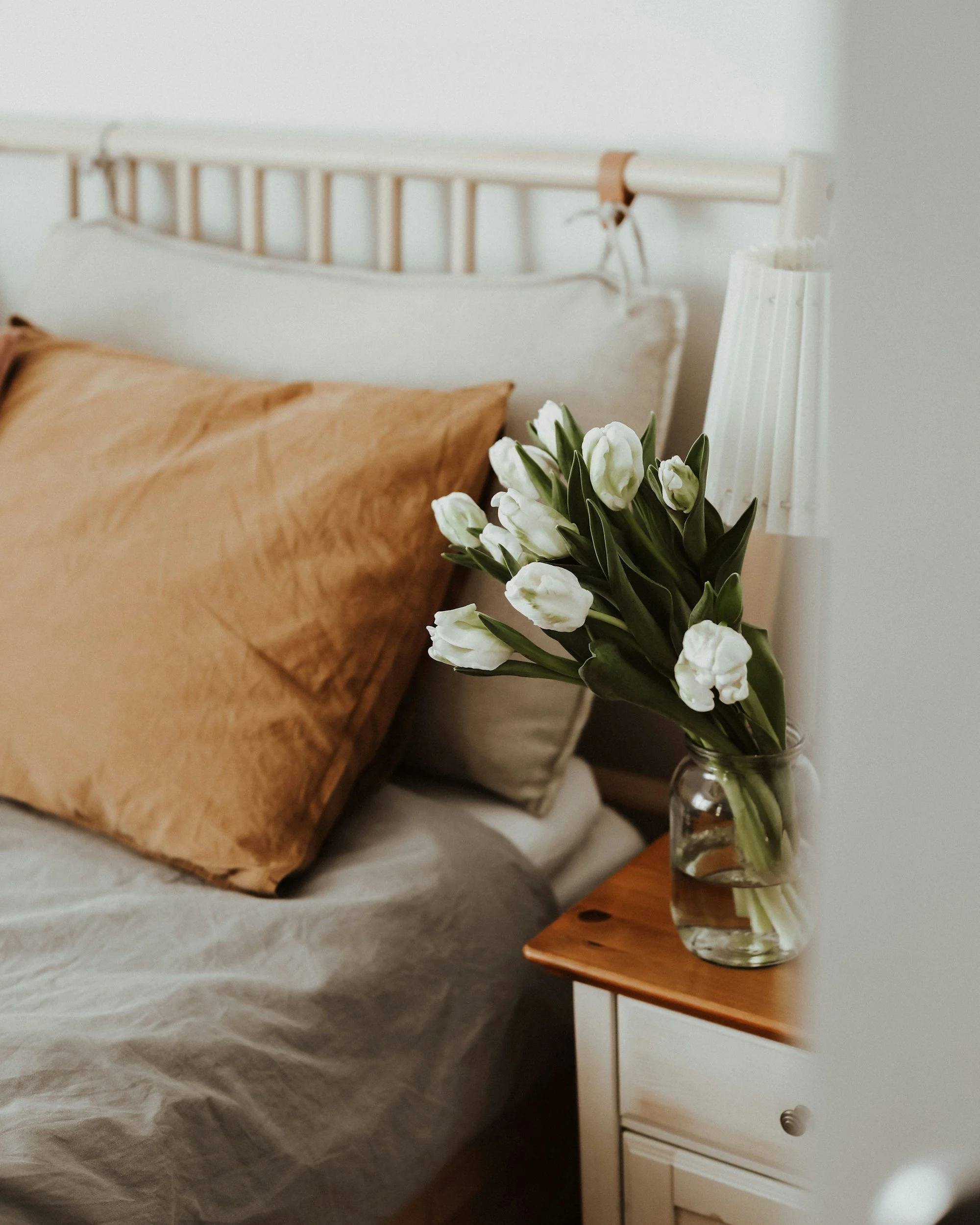 Close-up of a bed with a tan pillow, a white headboard, a nightstand with a glass vase of white tulips, and a white table lamp in a softly lit bedroom.