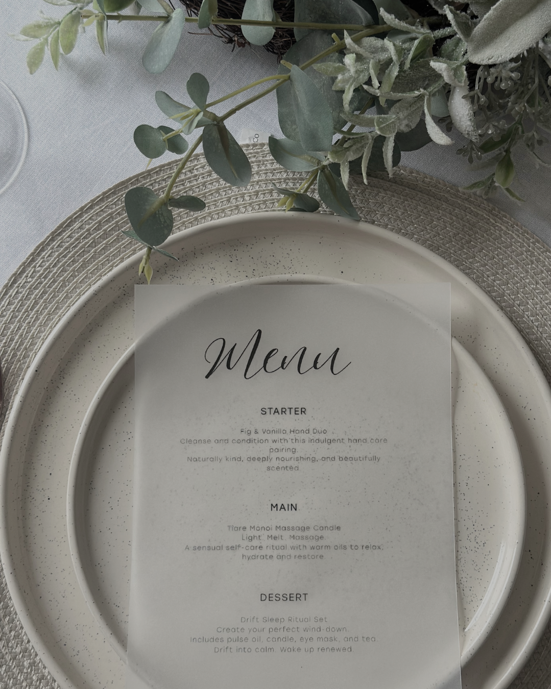 A menu placed on a white speckled plate with greenery and textured placemat underneath, set on a white tablecloth.