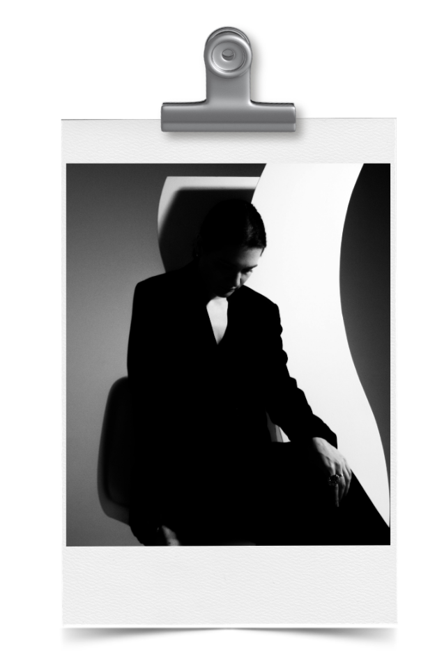 Black and white photo of a woman sitting on a chair against a background with abstract shapes, looking downward with her eyes closed, wearing a dark blazer.
