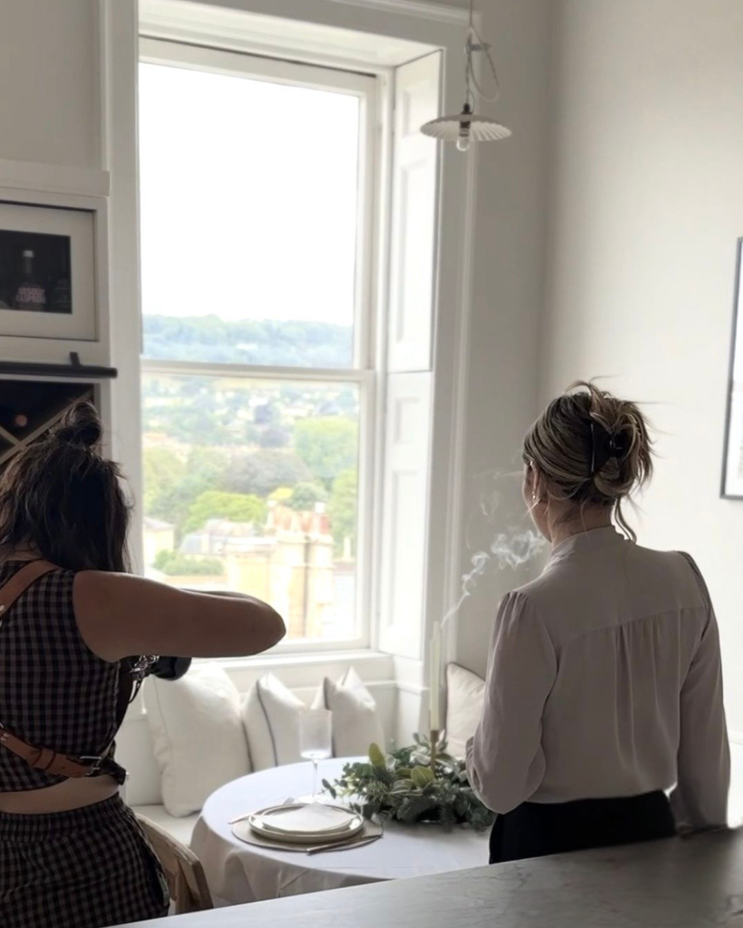 Two women inside a bright room near a large window, with one woman taking a photograph of the other who is standing by a dining table decorated with greenery, plates, and a glass of wine.