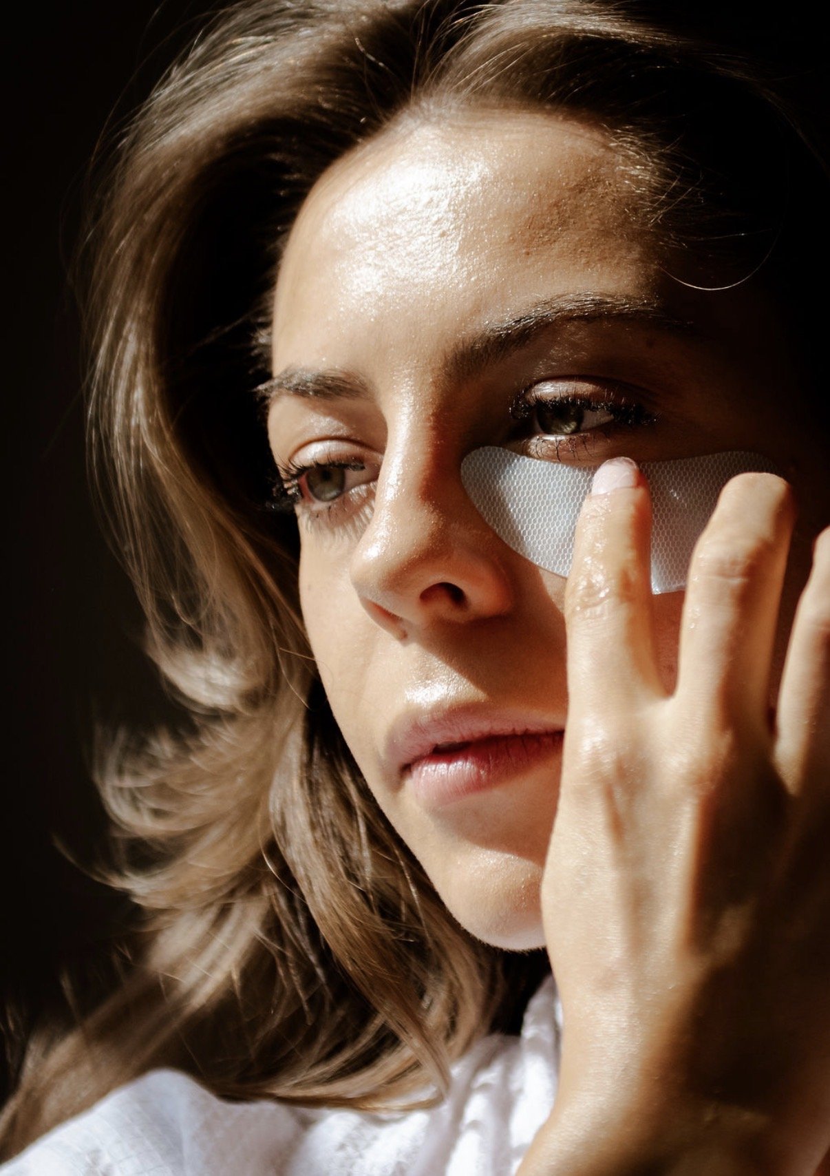 A woman with wavy hair applying a skincare patch under her eye.
