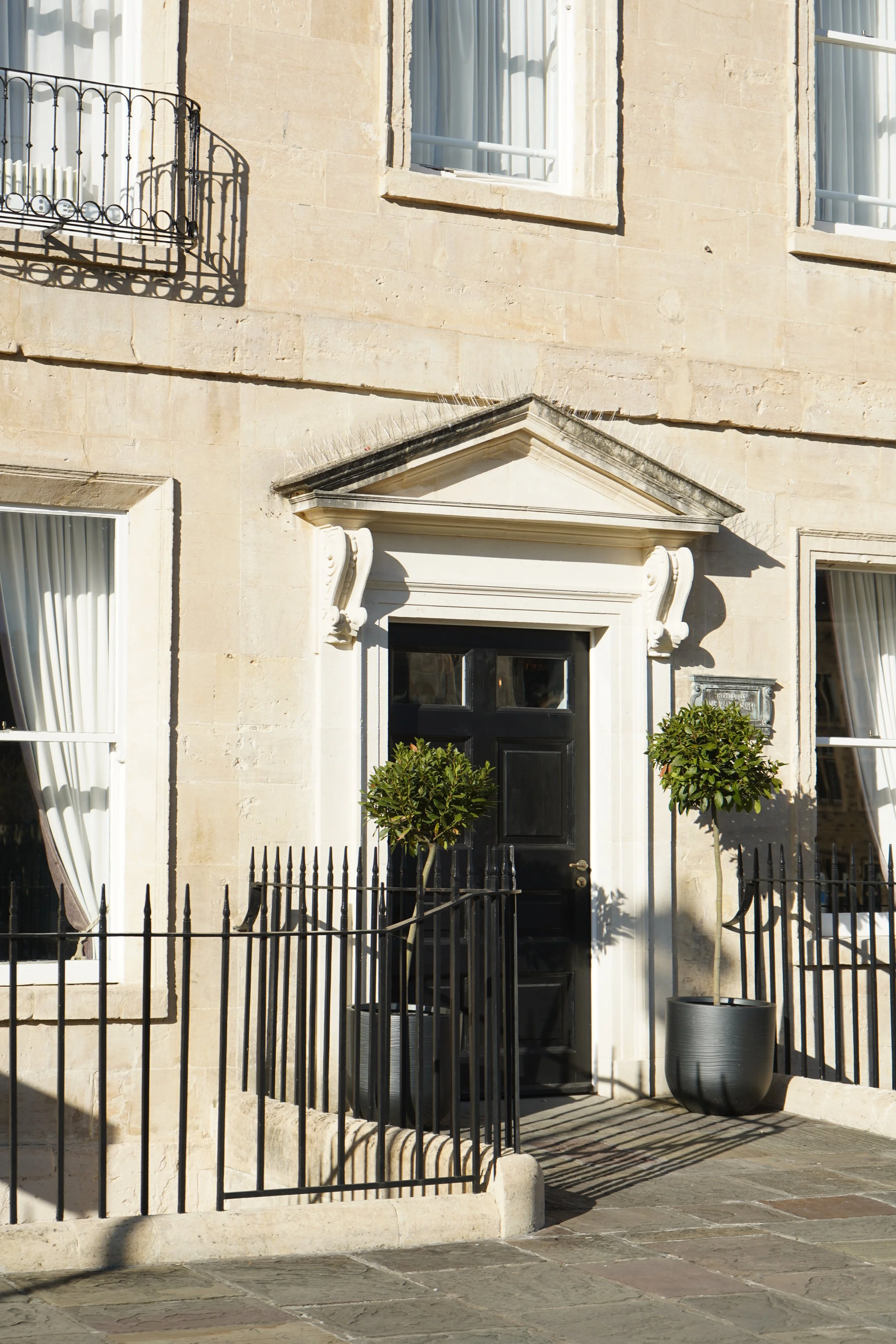Historic stone townhouse facade with black front door, ornate pediment, iron railings, windows, and potted shrubs