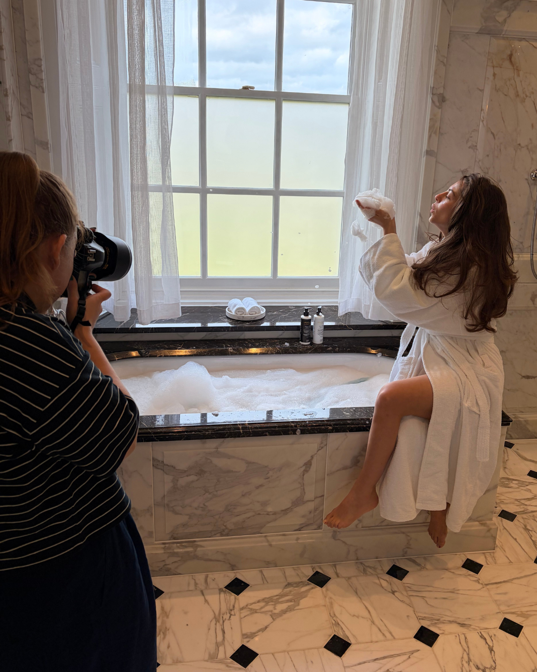 Woman in a bathrobe sitting on the edge of a bathtub, taking a shower or bath product while being photographed by another person in a striped shirt, in a luxurious bathroom with marble walls and black marble bathtub surround