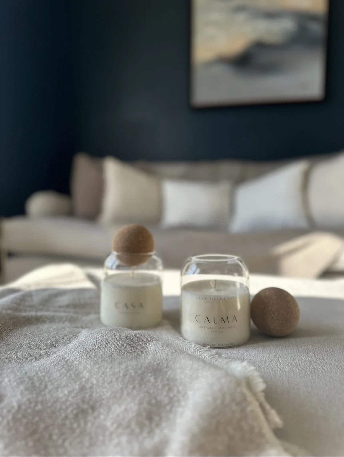 Two white candles labeled 'CALMA' with sand-colored decorative balls on top and beside them, placed on a light-colored fabric surface in a cozy room.