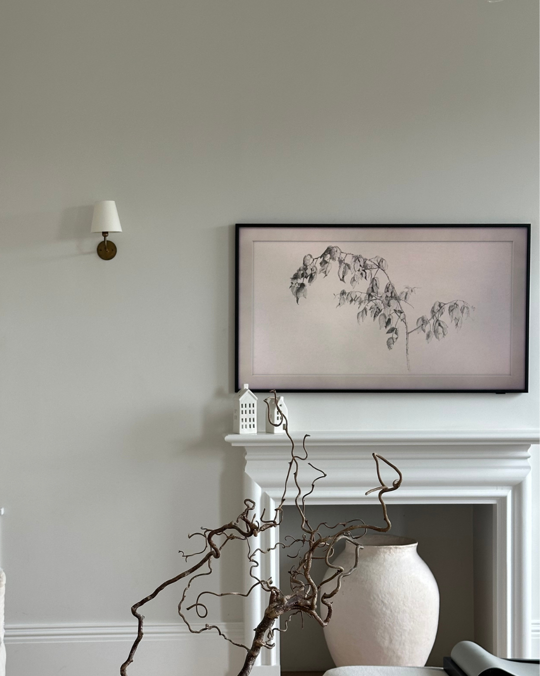 A living room corner with a white fireplace mantel, a large framed black and white botanical artwork hanging above, a ceramic vase inside the fireplace, a decorative white house model, a brown decorative branch, and a wall-mounted white and brass sconce.