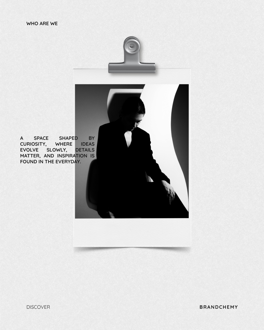 Black and white photo of a woman in a suit sitting in a uniquely-shaped space, with a close-up profile of a modern sculpture or architecture in the background. Text on the page includes 'Who Are We' at the top left, and a paragraph describing curiosity, ideas, details, and inspiration. The words 'Discover' and 'Brandchemy' are at the bottom.