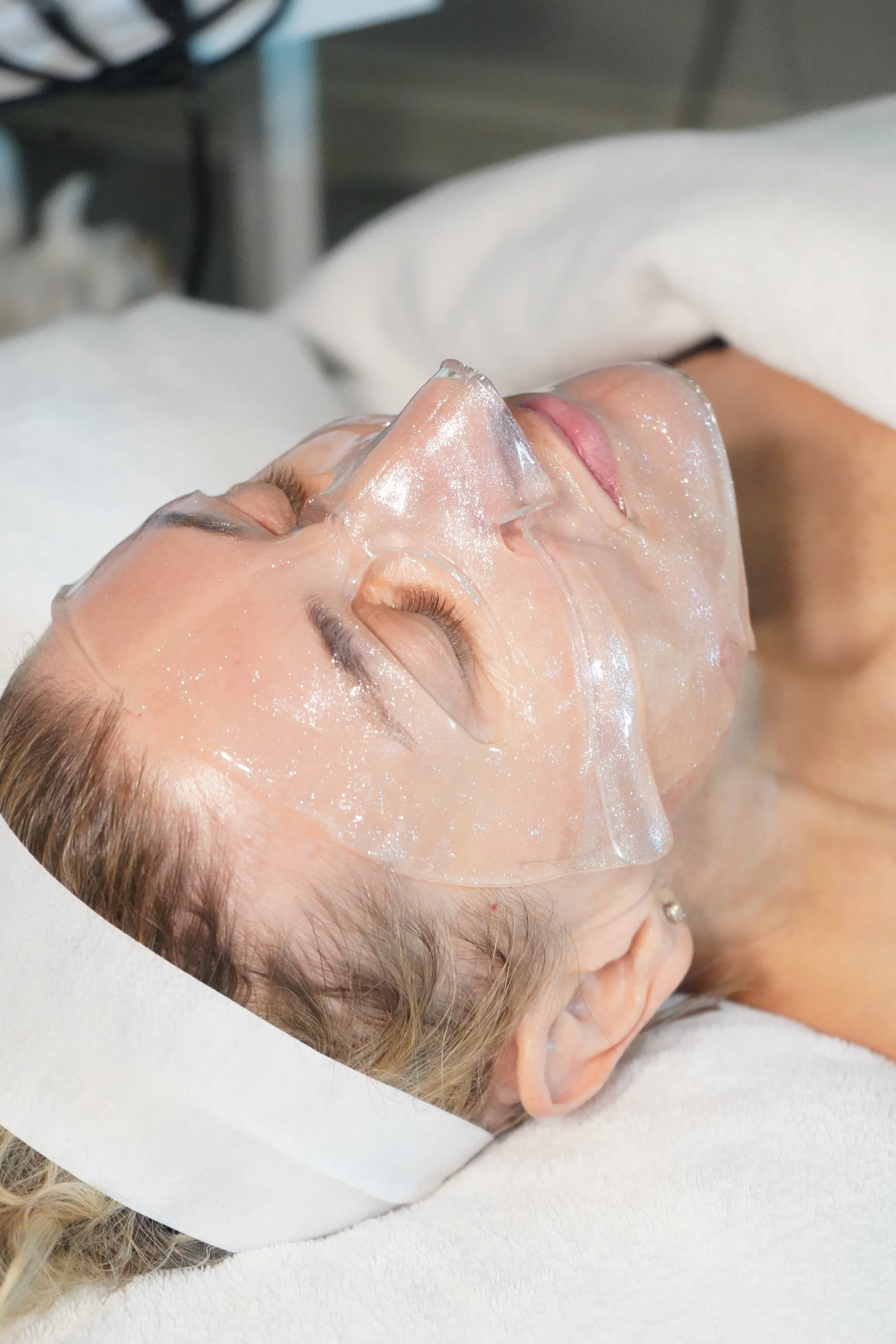Close-up of a woman receiving a facial treatment with a clear gel mask and protective eye covers.