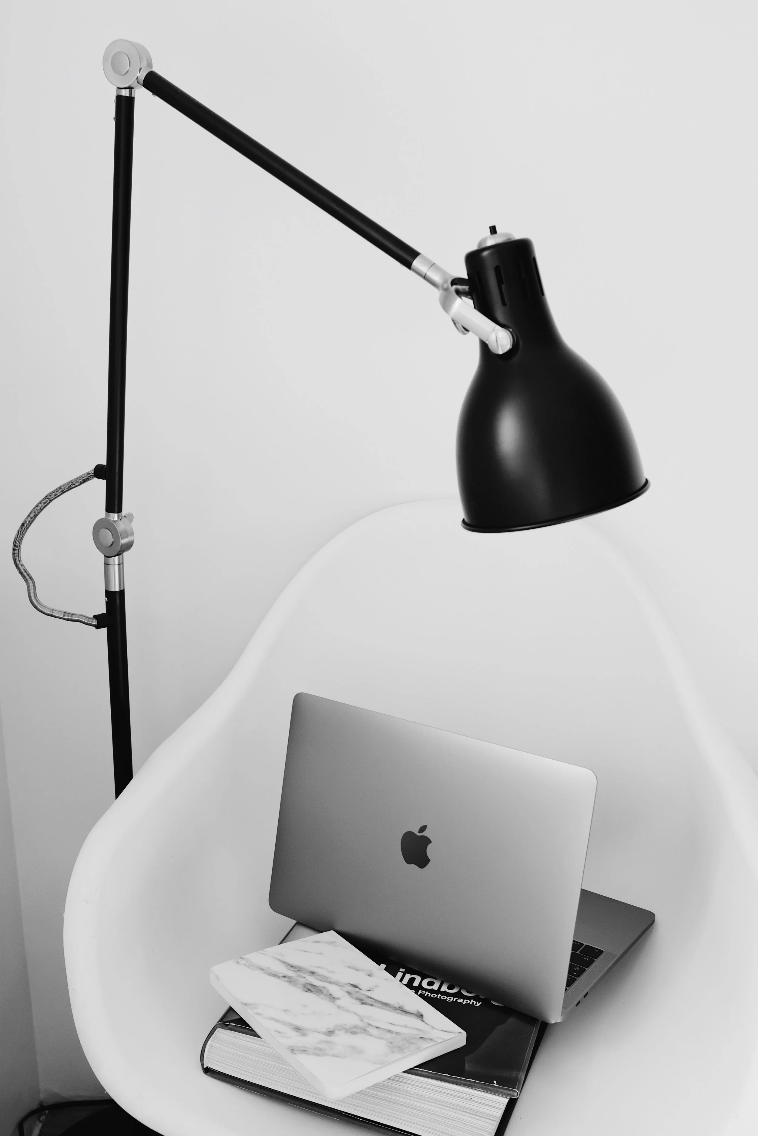 A workspace with a black desk lamp, an open MacBook, a marble-patterned notebook, and a closed book titled 'Kinab' on a white chair.