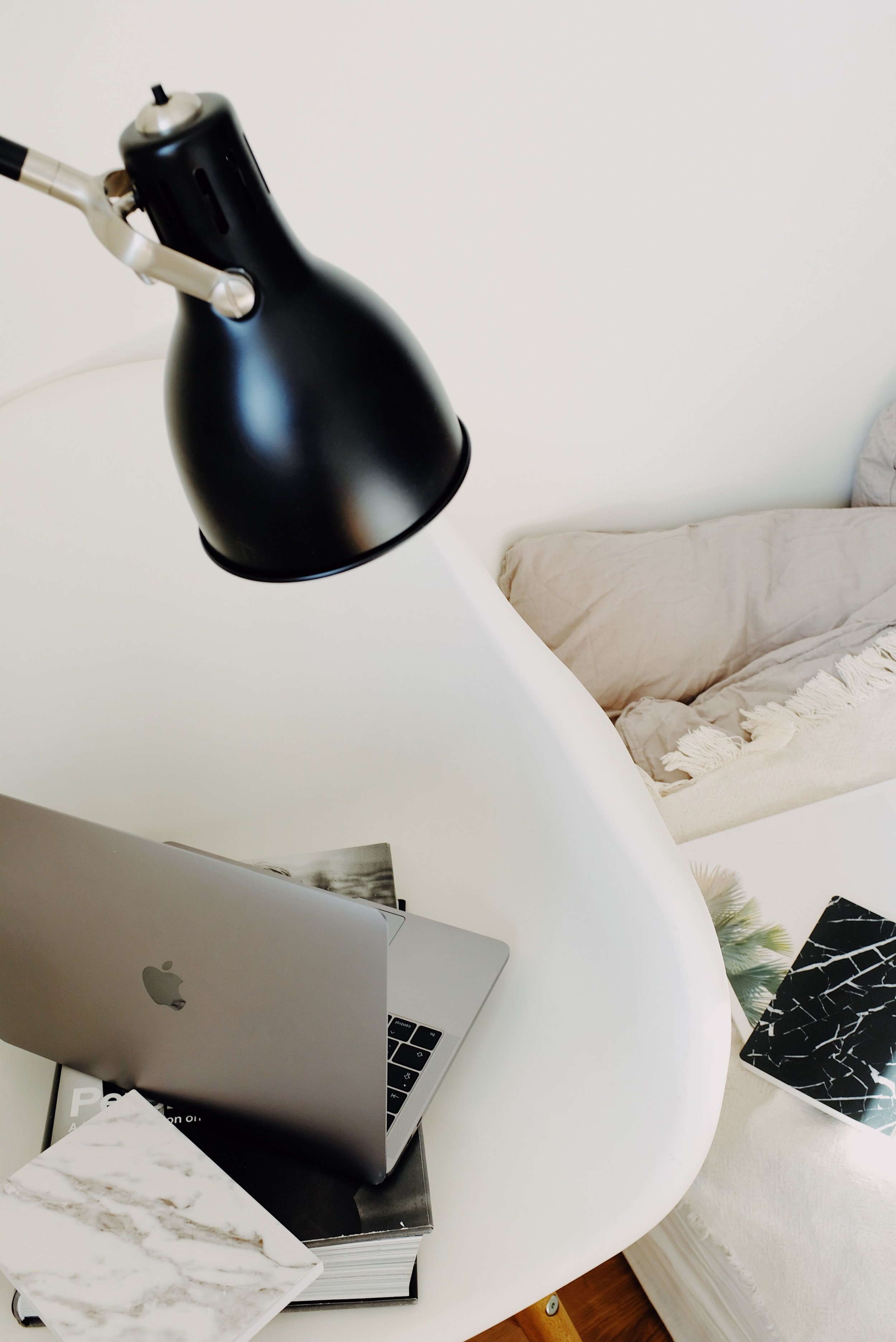 A workspace with an open MacBook, a marble tile sample, a coffee table book, a black and white patterned notebook, a silver desk lamp, and a beige couch with pillows in the background.