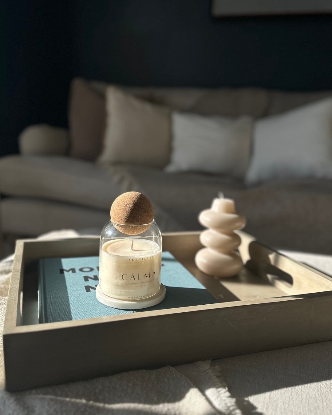 A tray with a lit candle labeled 'Calma' and a decorative white stack of stones, placed on a blue book, in front of a bed with pillows, bathed in natural sunlight.