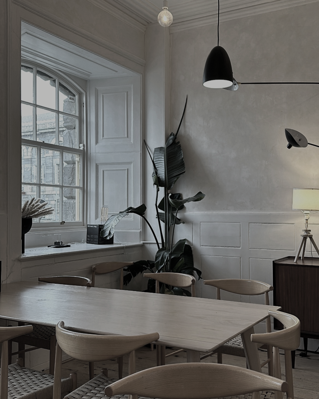 Interior of a modern dining or meeting room featuring a large wooden table, light wood chairs, a plant near a window, a dark wooden cabinet with a lamp, and black hanging pendant lights.