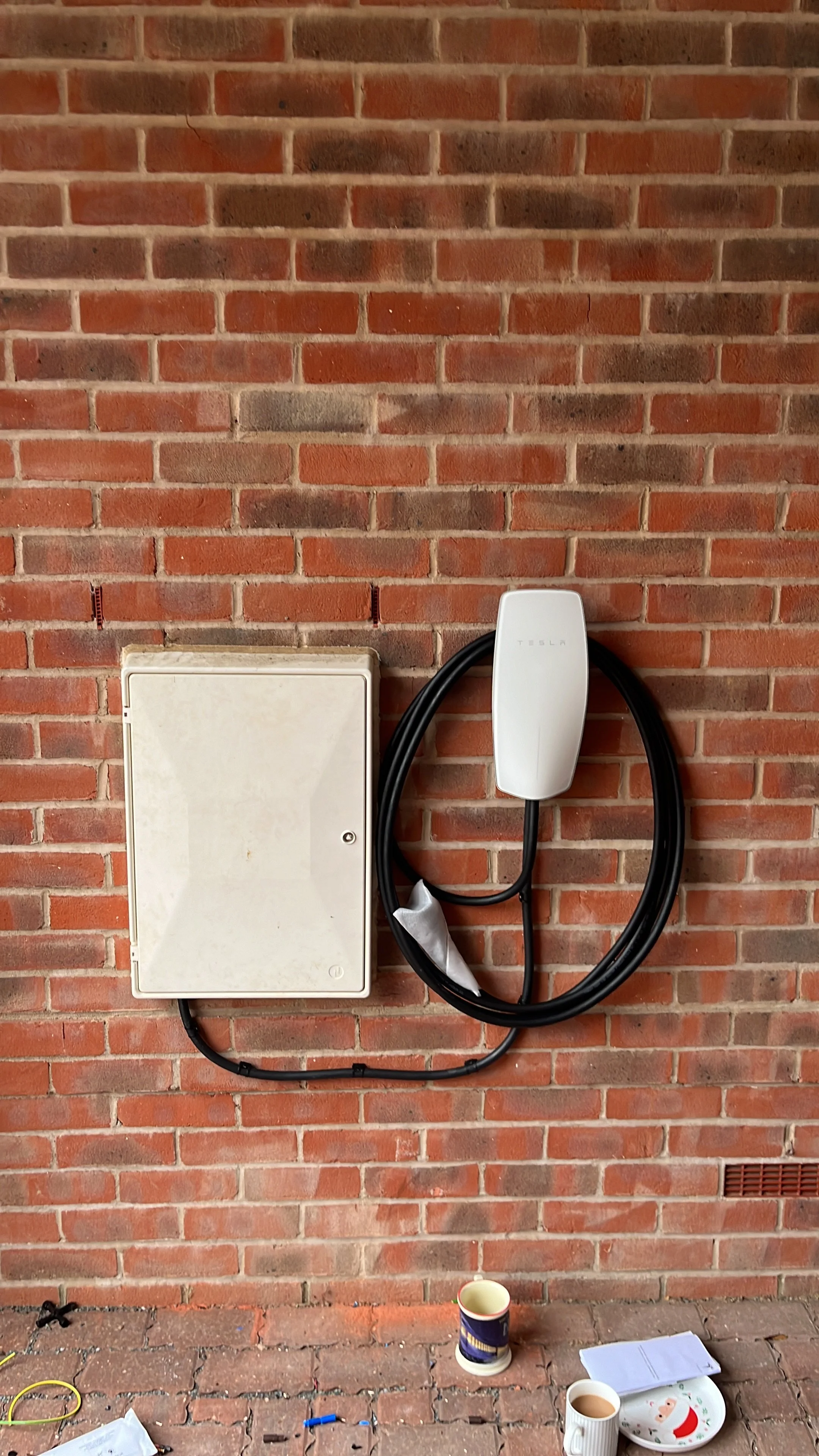 Electric vehicle charging station mounted on a brick wall with an electric charging cable hanging down, and a small table with cups, papers, and a decorative Santa plate in front of the wall.