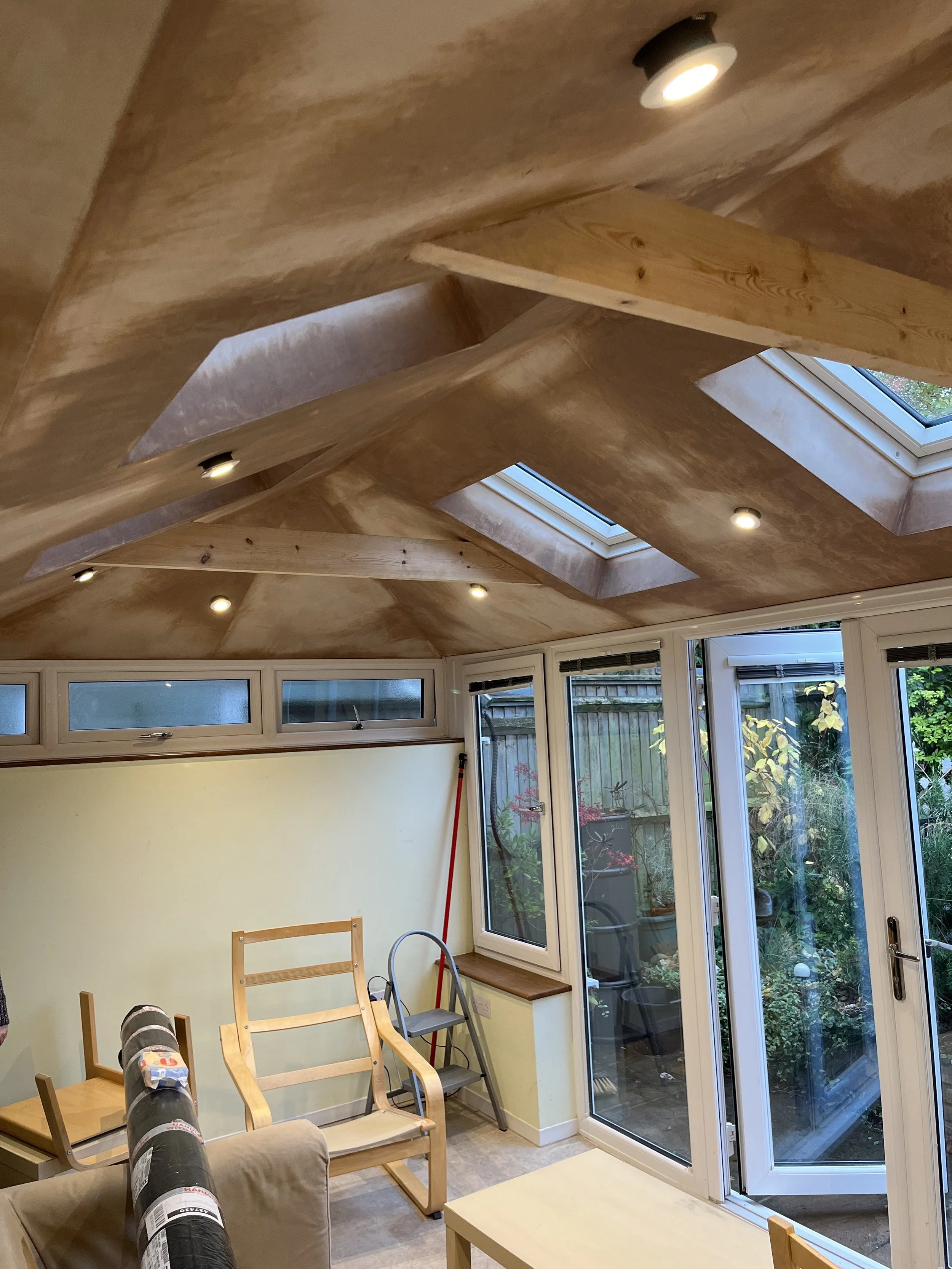 Living room with a ceiling equipped with two skylight windows and illuminated by recessed lighting. The room has large glass doors opening to a garden, with blooming bushes outside. The room contains a partially assembled wooden chair, a sofa, a dining table, and other furniture items, with cleaning tools visible in the corner.