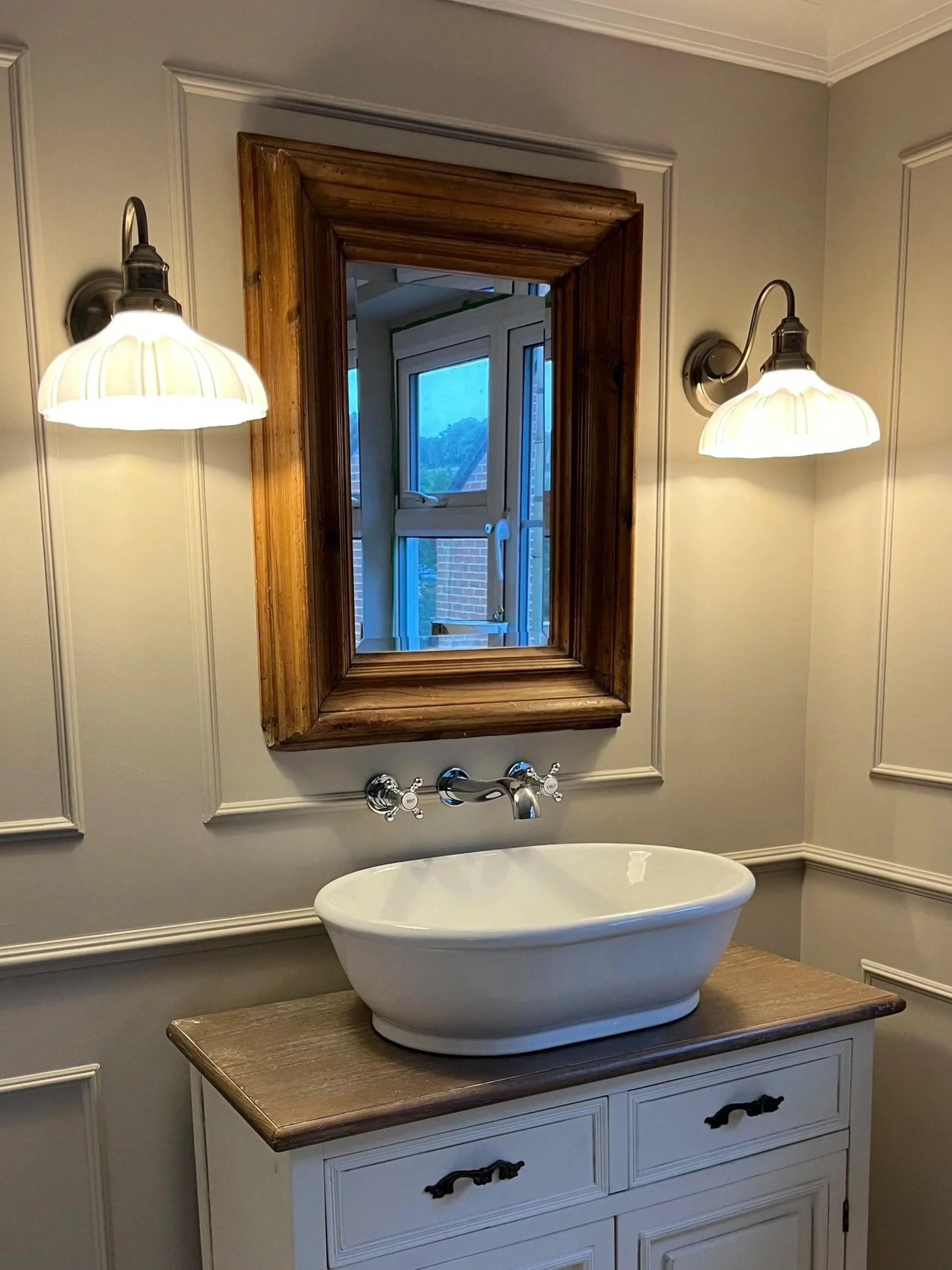 Bathroom with a white bathtub, a wooden framed mirror above the sink, and two wall-mounted light fixtures with white shades.