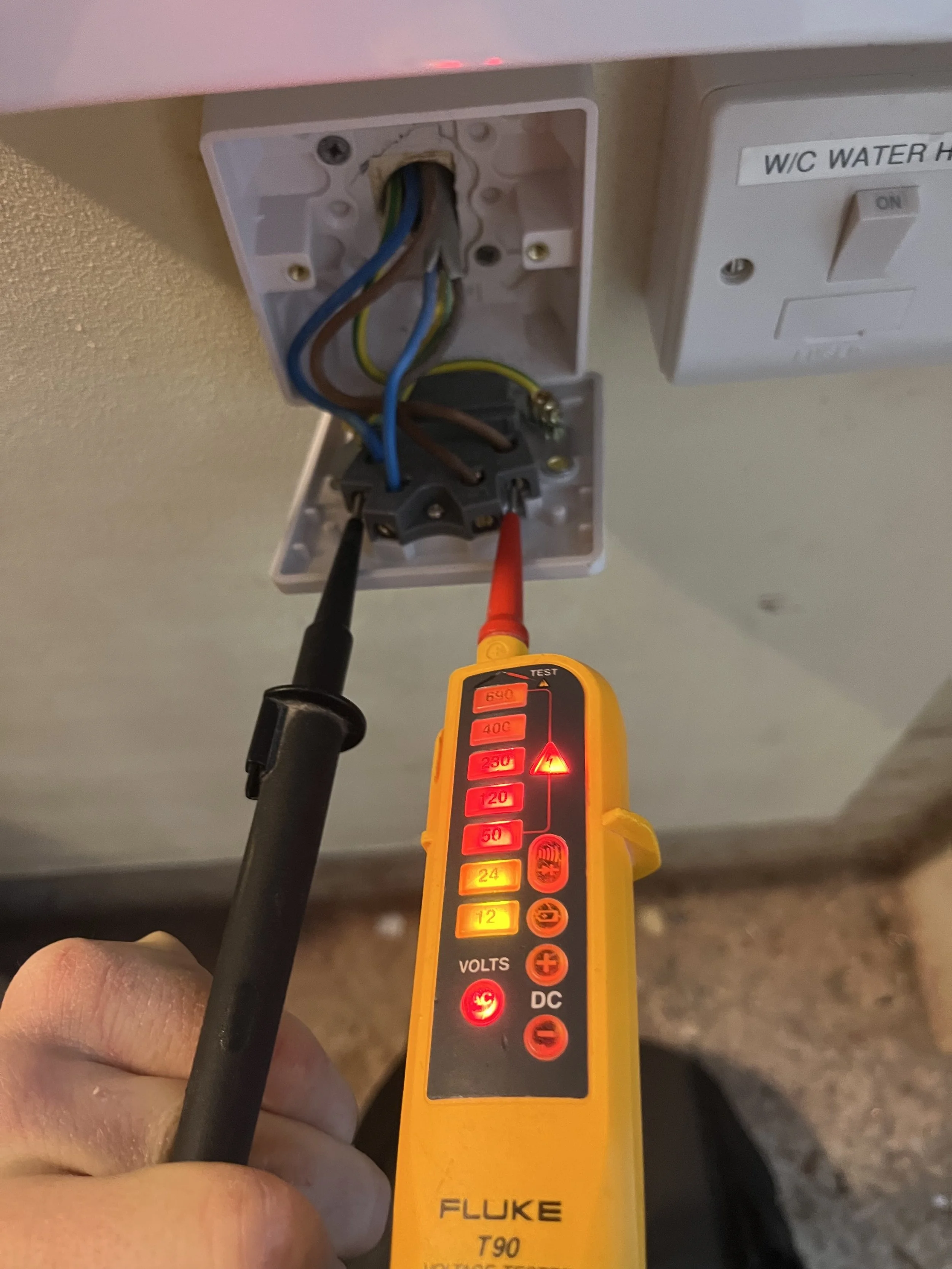 A person's hand holding a multimeter testing electrical wires inside an open electrical outlet box on a ceiling. The outlet box contains several colored wires, and the multimeter displays a reading of 640 volts.