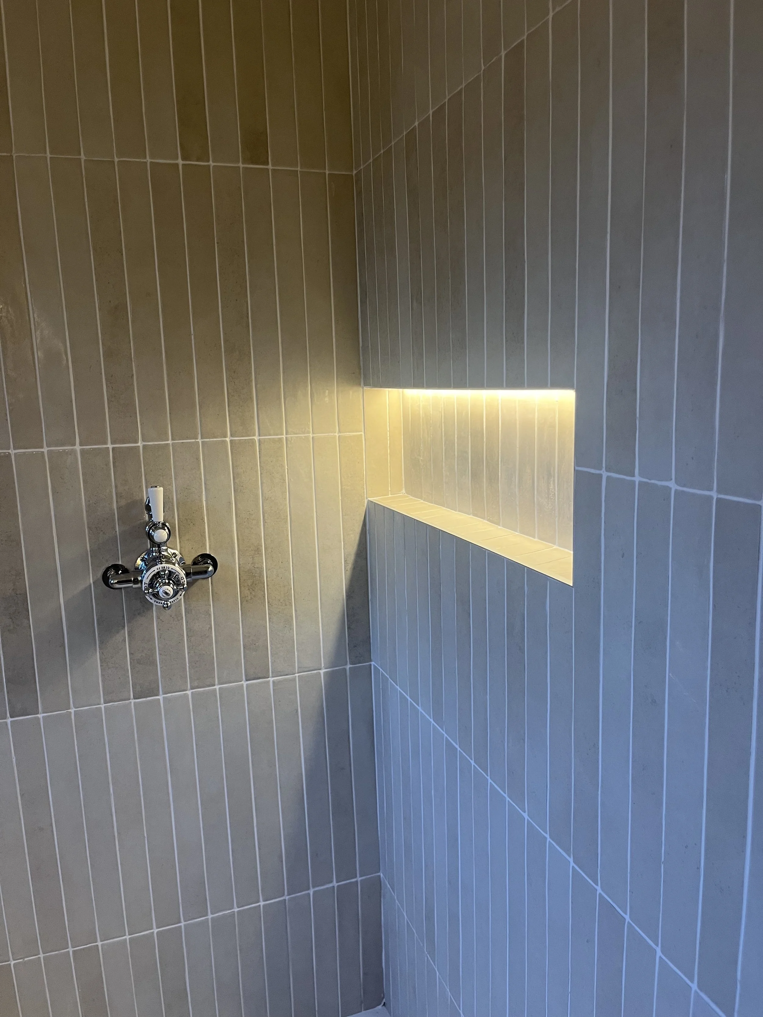 A bathroom shower wall with beige tiles, a built-in shelf with LED lighting, and a chrome shower valve.