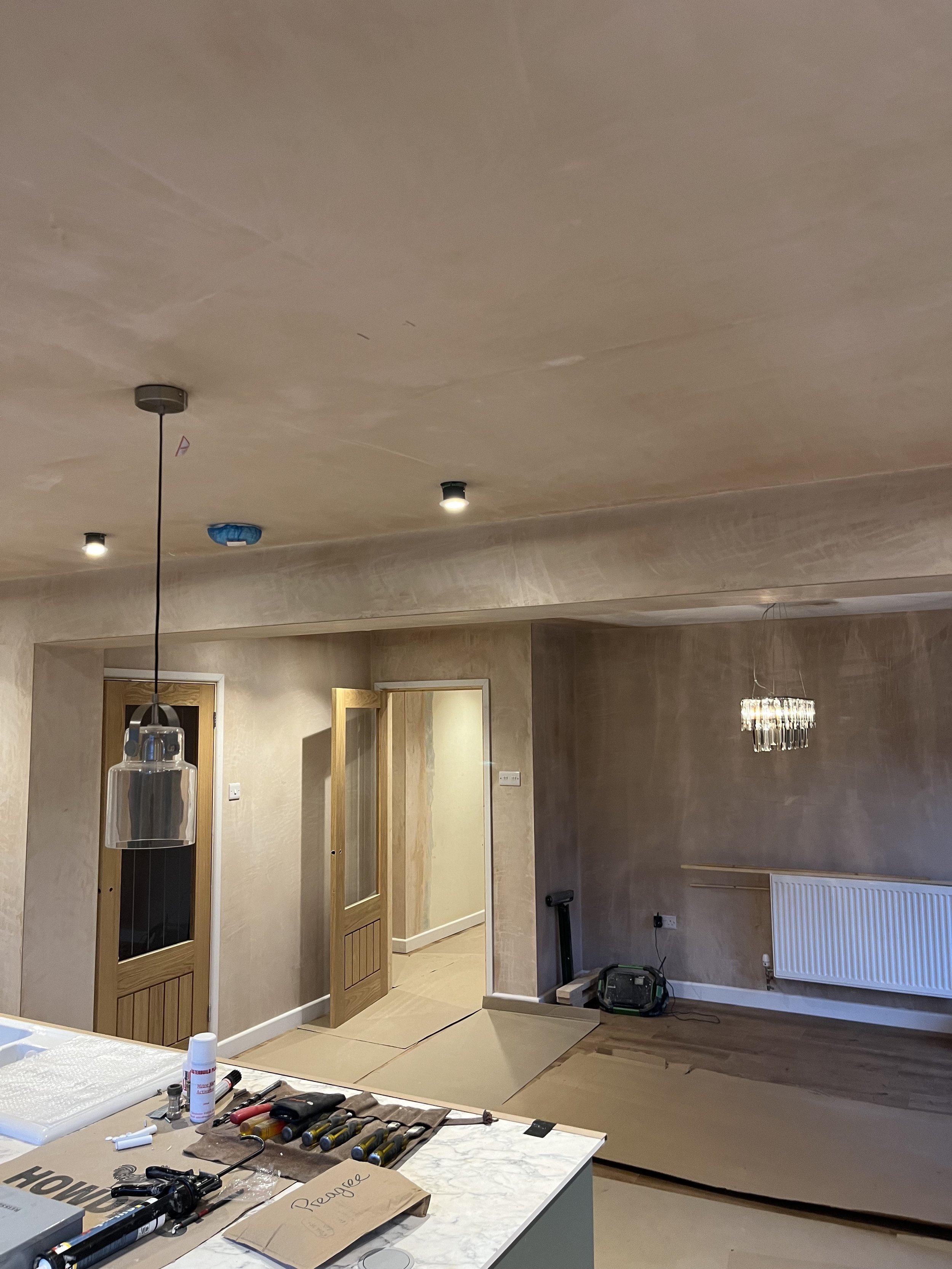 Interior view of a room under renovation with unfinished ceiling, open doorway, construction tools on a counter, and tools and materials on the floor. A chandelier hangs from the ceiling and a radiator is installed on the wall.