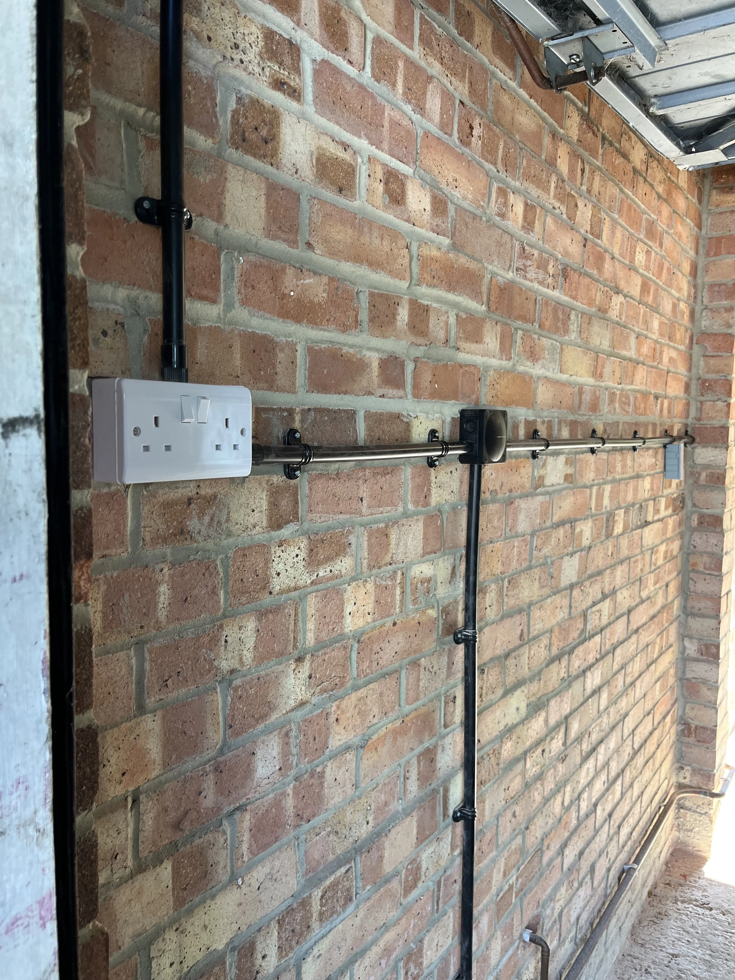 Close-up of a brick wall with electrical wiring and a metal conduit pipe running along it, connected to an electrical outlet box.