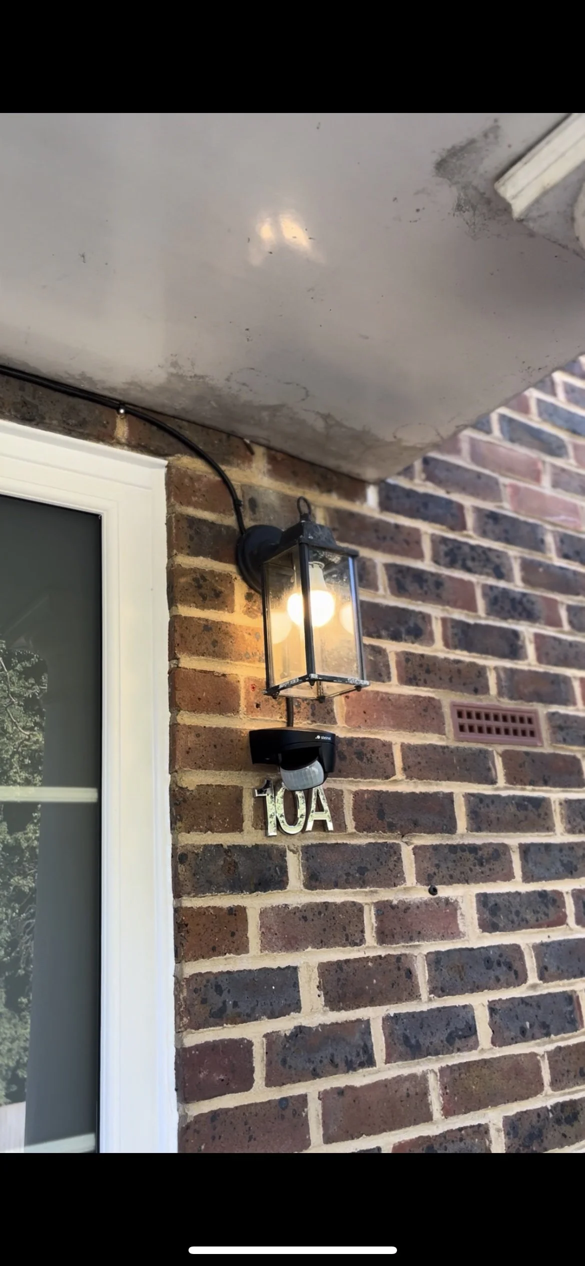 Exterior brick wall with a house number 10A and a black outdoor lantern light fixture, with a motion sensor below the light.