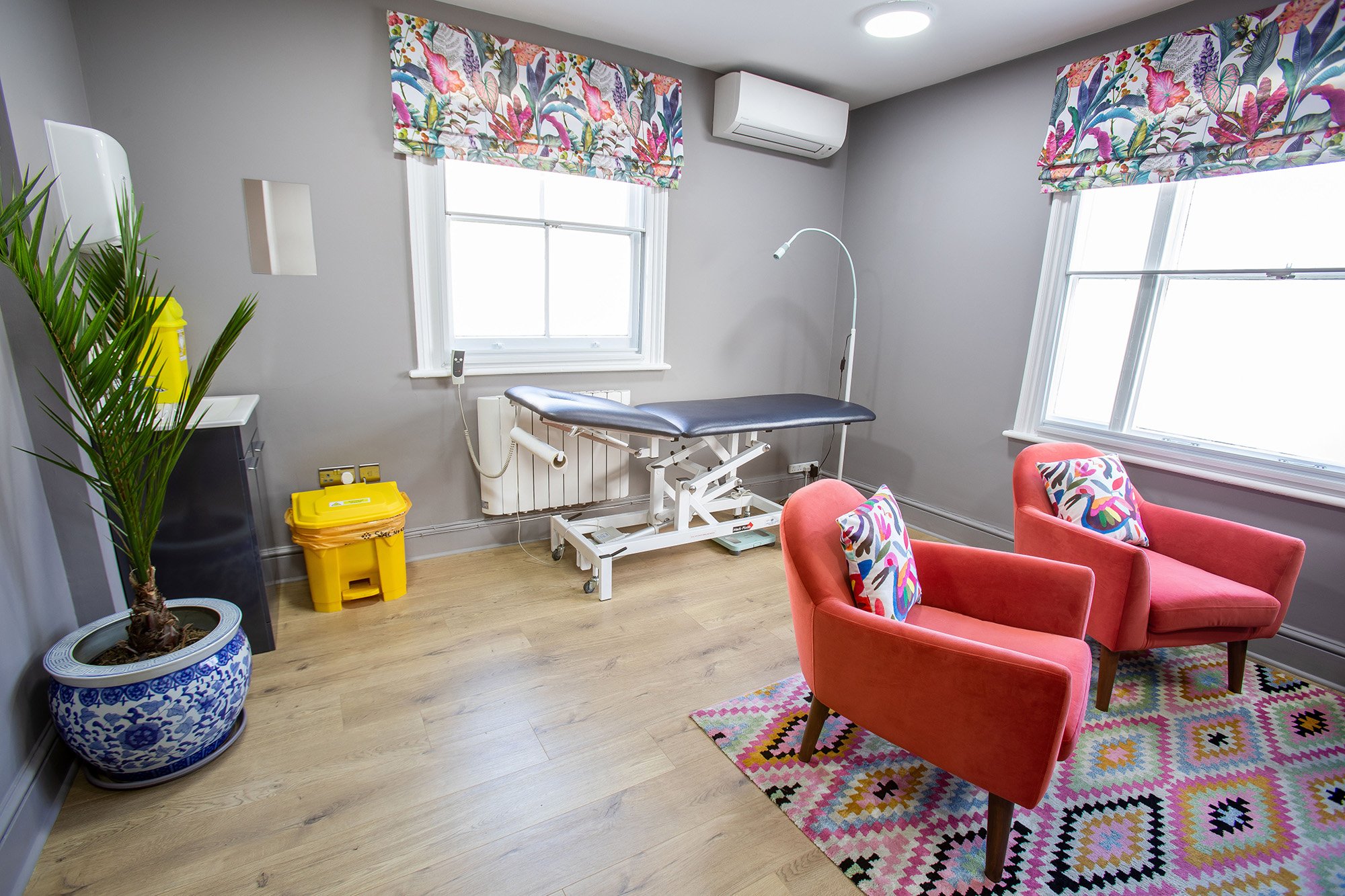 Interior view of a medical examination room with an examination bed, colourful chairs with patterned cushions, a potted plant, and windows with floral curtains.