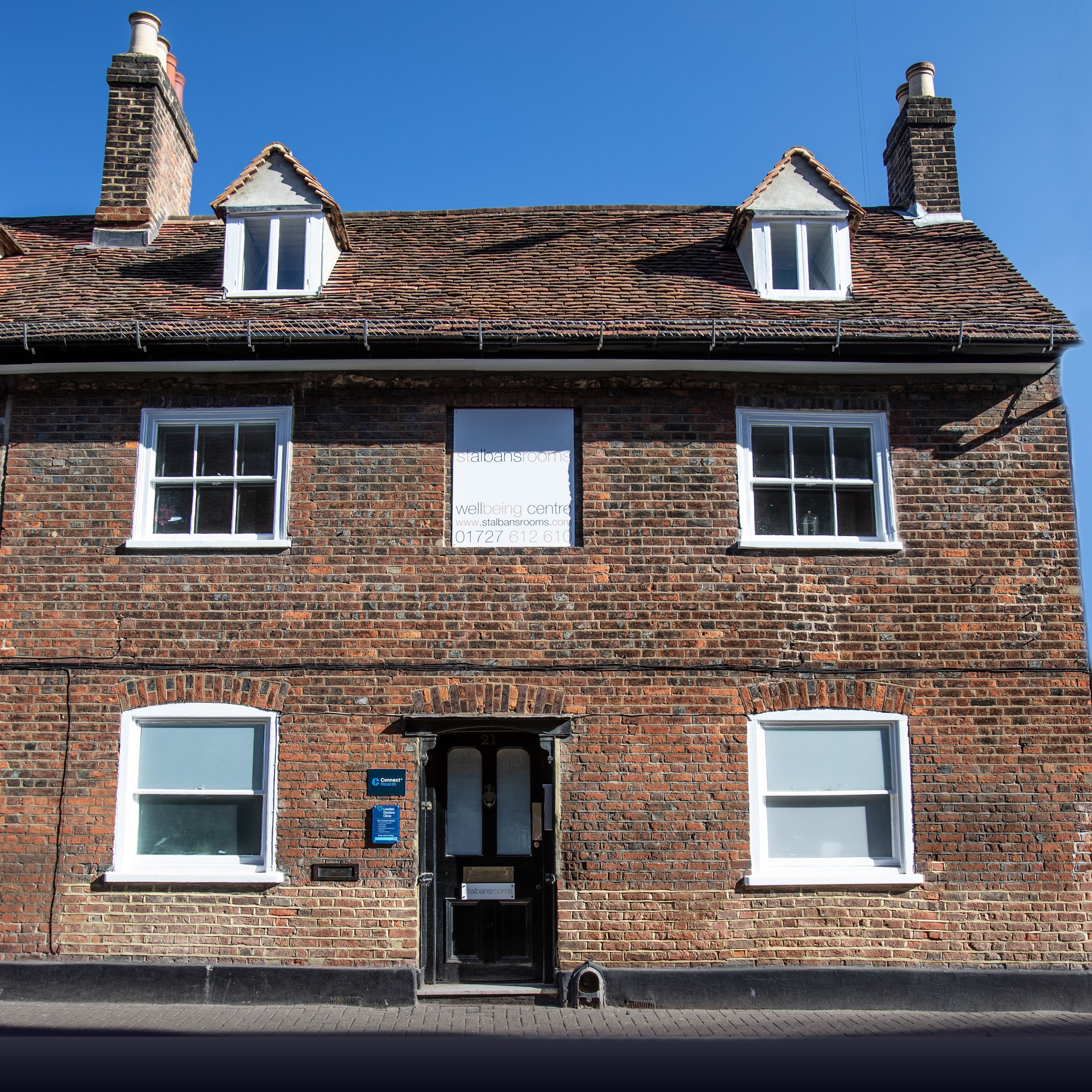 Brick building with two white-framed windows on either side of a black front door, two dormer windows on the roof, and a sign on the door reading 'Stalbans rooms' with contact information below.