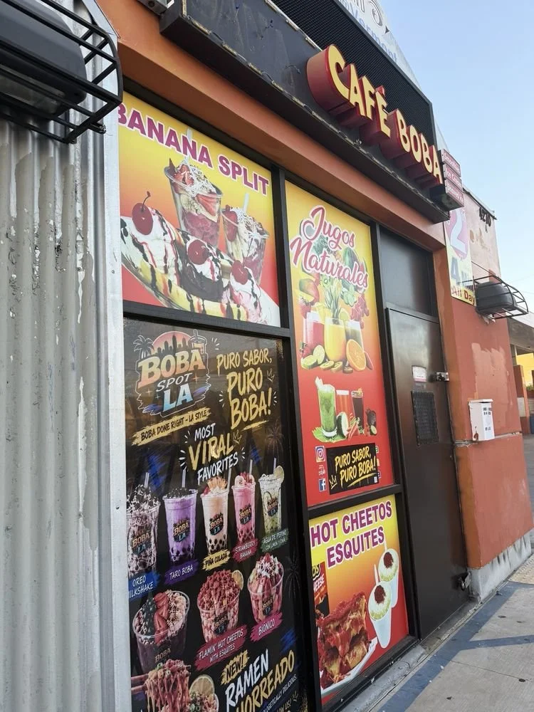 Signage for Cafe Boba, featuring images of banana split ice cream, natural juices, hot cheetos, and esquetes, along with promotional posters for bubble tea flavors and ramen.