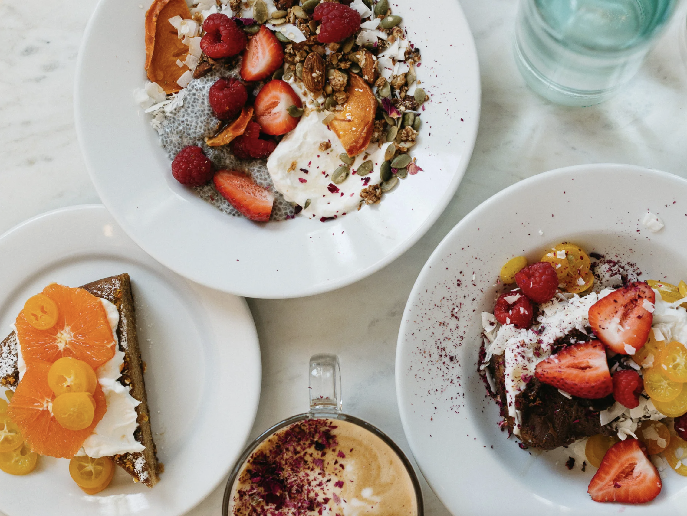 A top view of a white plate with assorted desserts, including a piece of cake with orange slices and yellow cherry toppings, a plate of yogurt topped with raspberries, strawberries, pumpkin seeds, granola, and coconut flakes, and other colorful desse