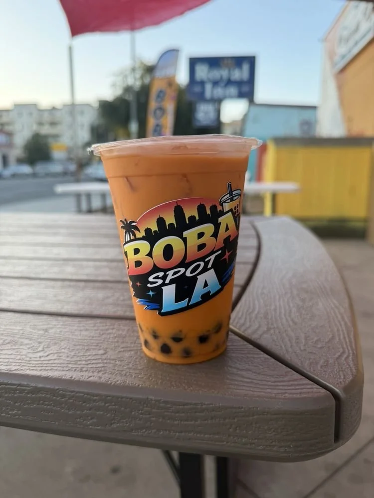 A cup of bubble tea with black tapioca pearls at the bottom, sitting on a wooden outdoor table. The cup has a colorful logo that says "BOBA SPOT LA" with a silhouette of the Los Angeles skyline and palm trees.